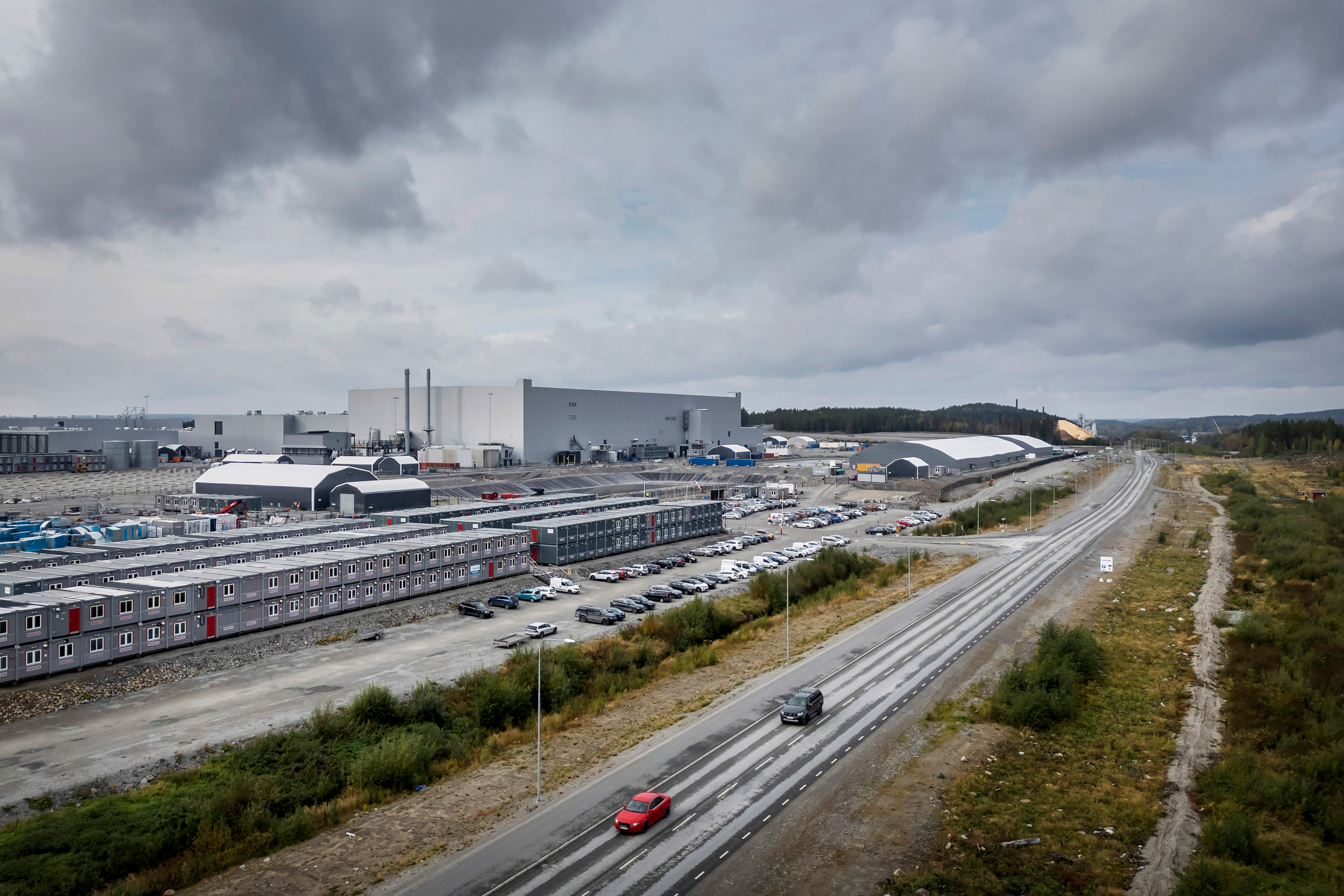 Luftbild av ett industriområde med stora byggnader, parkeringsplats fylld med bilar och intilliggande vägar. Molnigt himmel i bakgrunden.