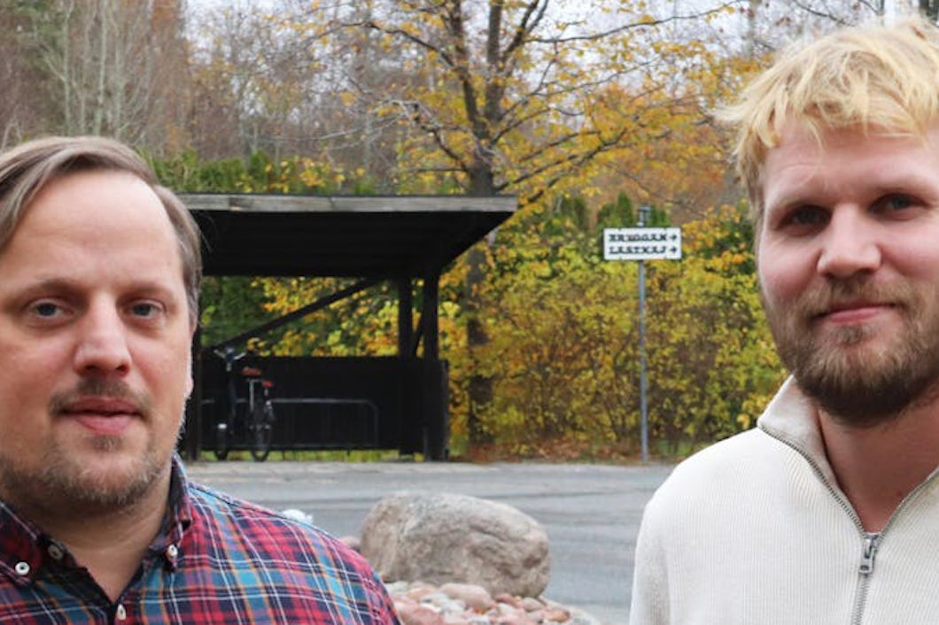 Två män står utomhus i en park under hösten. Den ena har en rutig skjorta och den andra bär en vit tröja. I bakgrunden syns träd och bänkar.