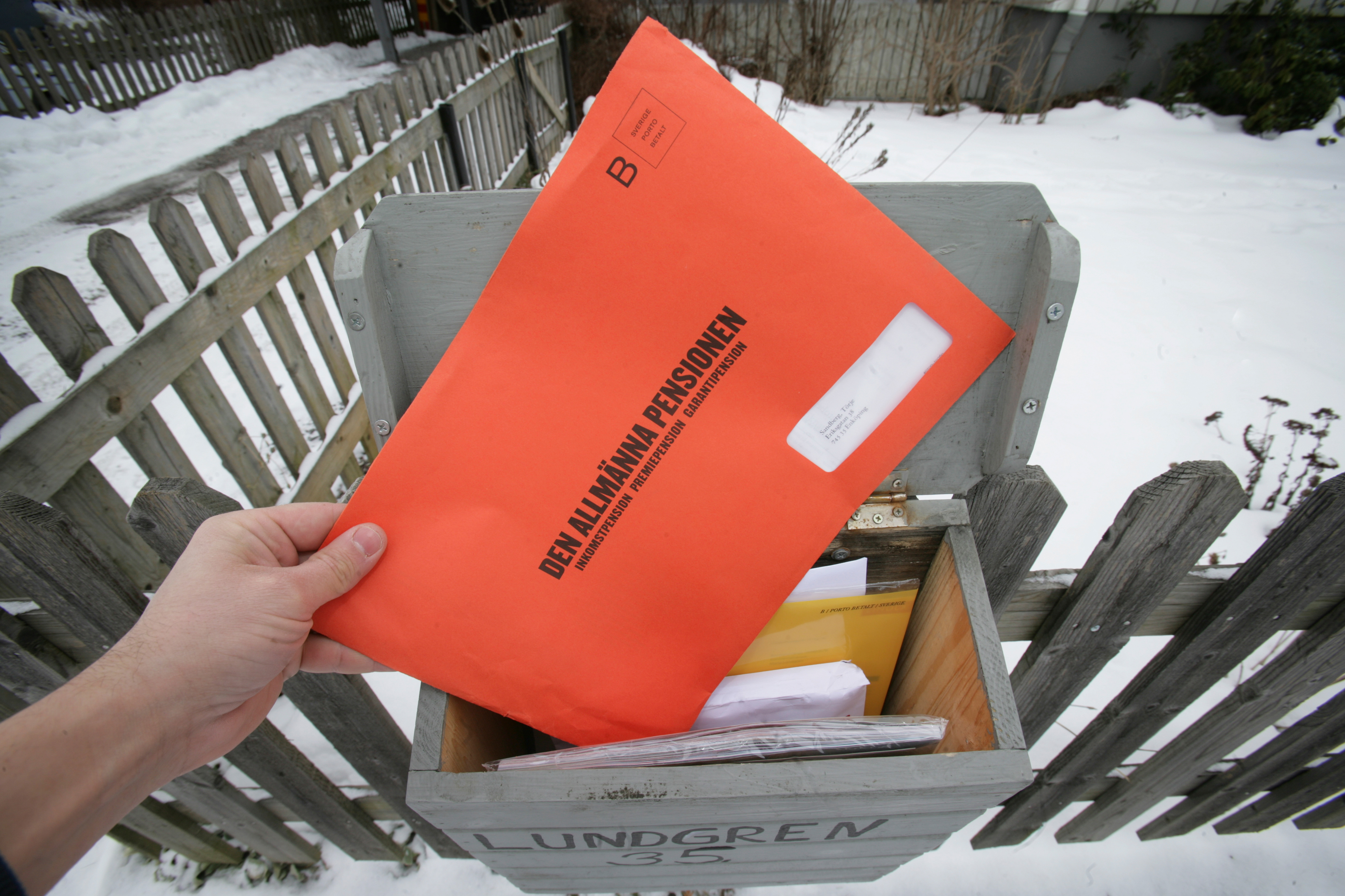 En hand håller ett orange kuvert märkt "Det Allmänna Pensionen" ovanför en brevlåda fylld med brev. I bakgrunden syns snö.