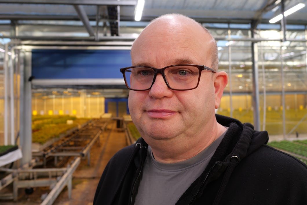 En man med glasögon står i ett växthus omgiven av växter.