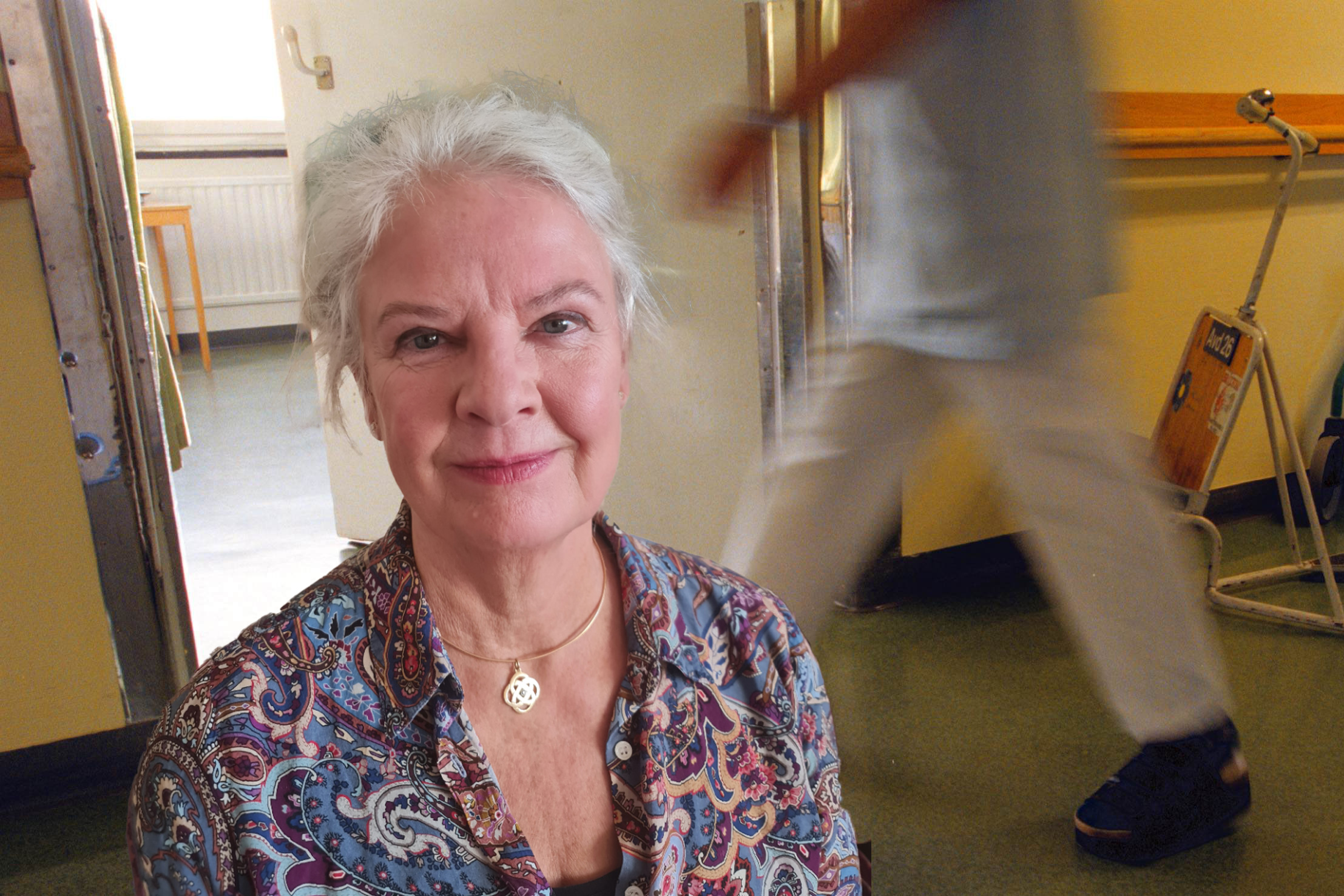 En äldre kvinna med vitt hår och en mönstrad blus sitter i ett rum och ler medan en person går förbi i bakgrunden.