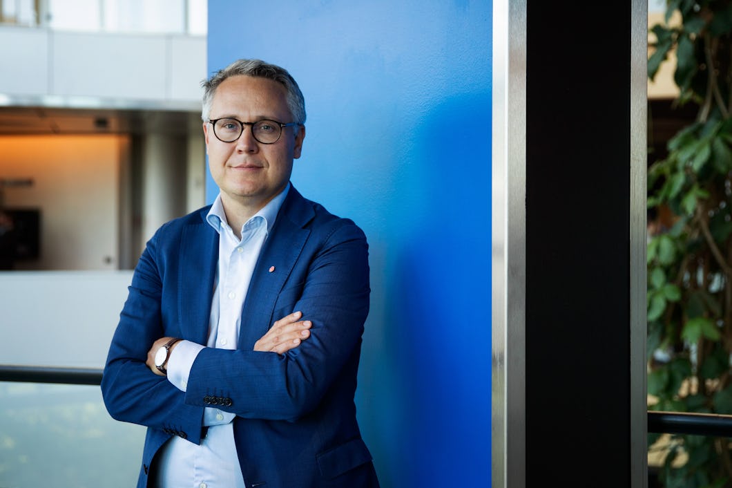 Person i blå kostym och glasögon står med armarna i kors bredvid en blå vägg.