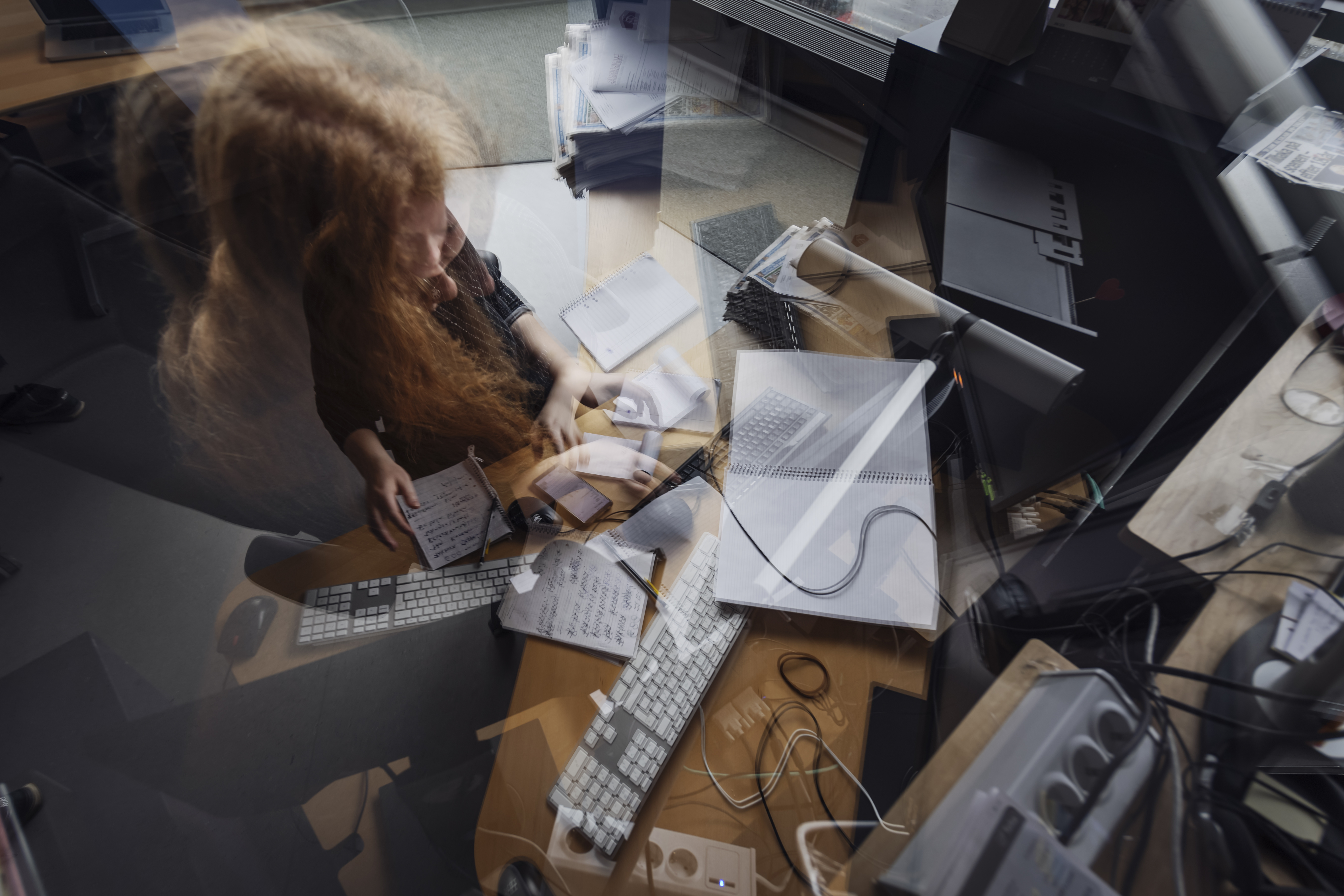 Översiktsbild av ett skrivbord fyllt med papper, datorer och olika kontorsmaterial. En person med långt hår skriver anteckningar omgiven av kablar och elektroniska enheter.