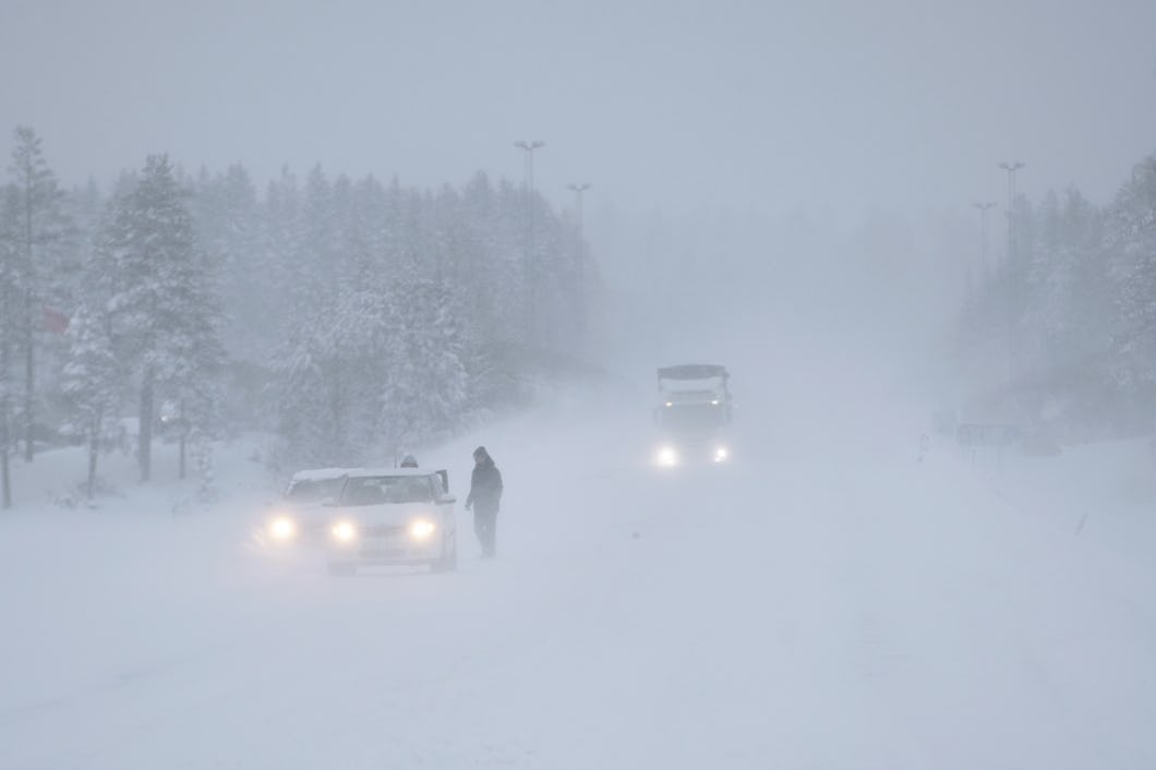 En person står bredvid en bil på en väg täckt av snö, där det snöar kraftigt.