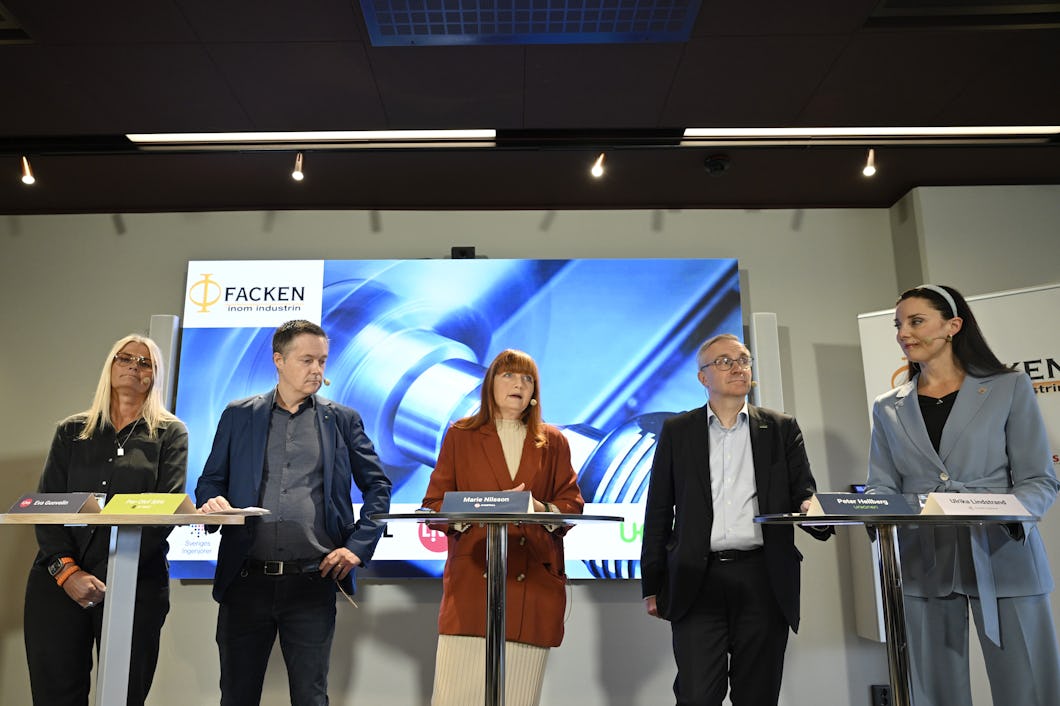 Fem personer står vid talarpodium i en konferenslokal. Bakom dem finns en stor skärm som visar bilder av industriella maskiner.