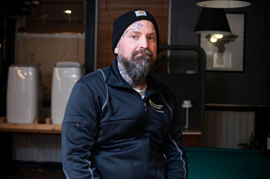 Stefan Fernaeus med skägg och tatueringar sitter på ett biljardbord, iklädd en svart jacka, mössa och slitna jeans i ett svagt upplyst rum.