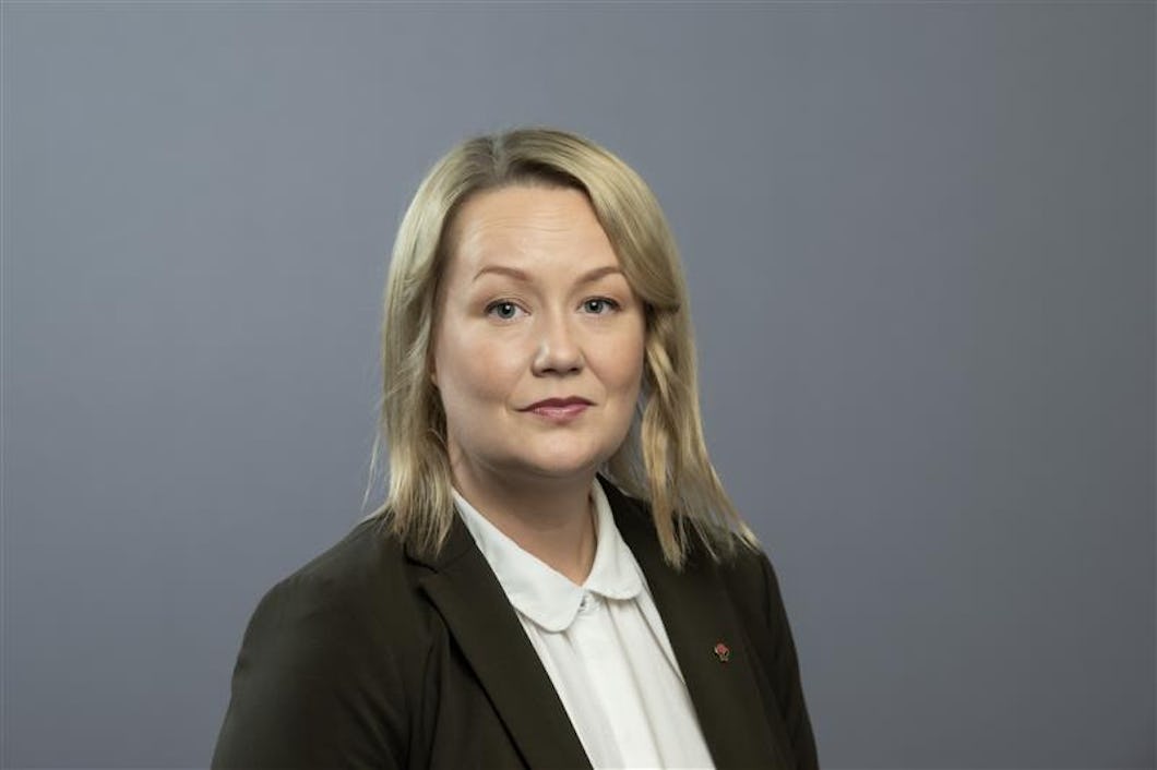 Stephanie Bruksgård med axellångt blont hår, iklädd en mörk kavaj och vit skjorta, står mot en enkel grå bakgrund.