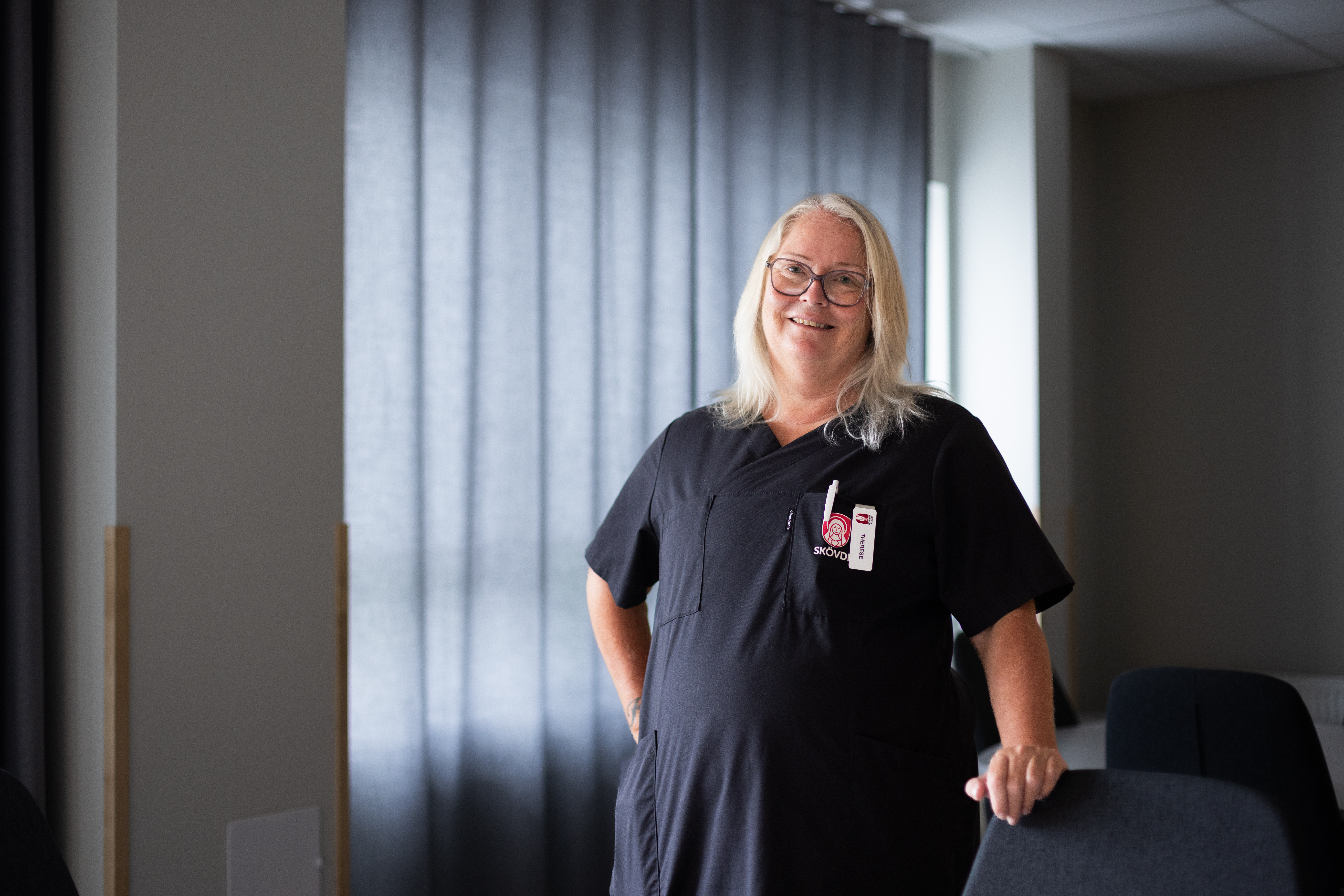 En person med långt hår och glasögon står i ett rum med grå gardiner, iförd en svart uniform.