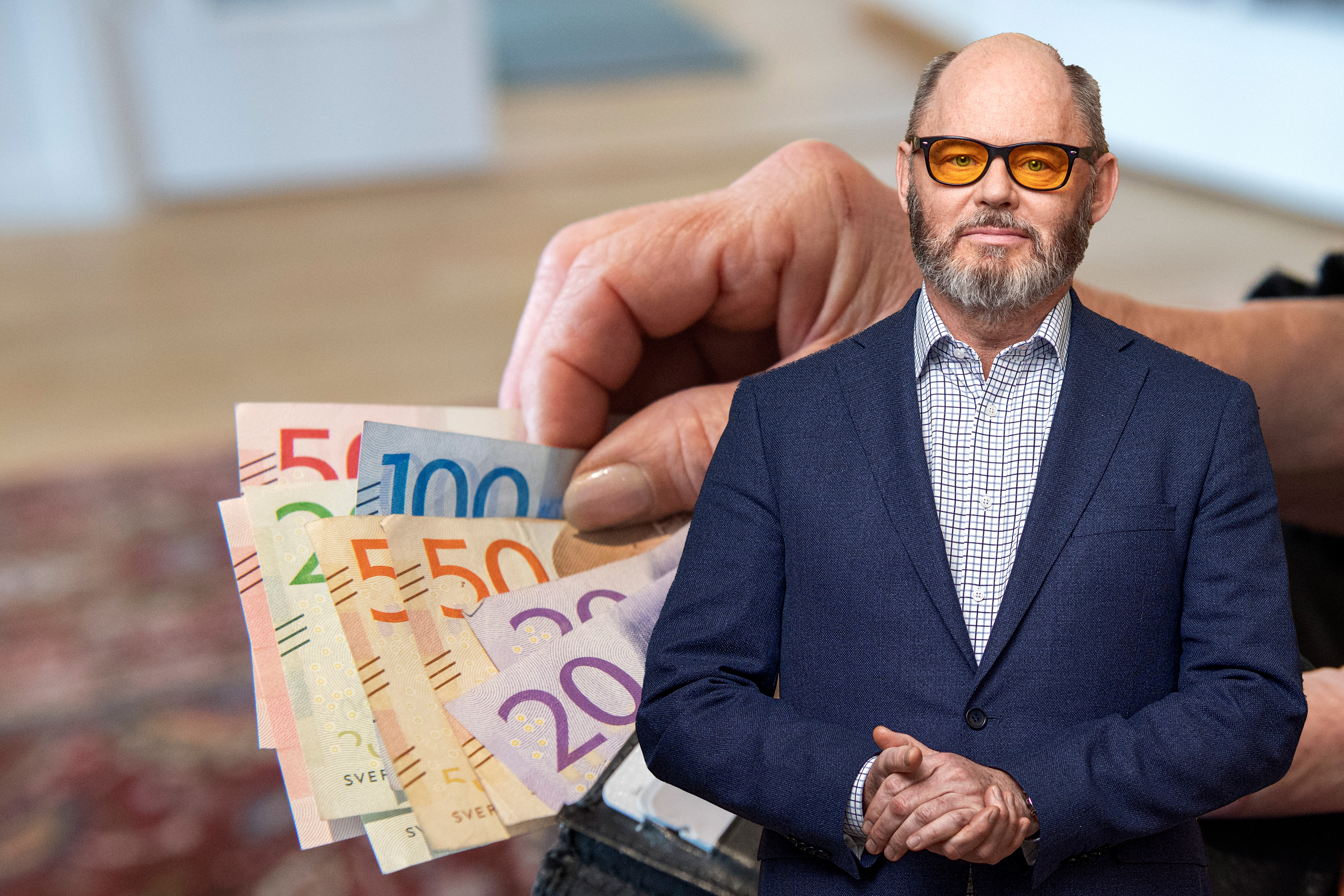 En person håller svenska sedlar med en man i kostym och glasögon överlagd framför.