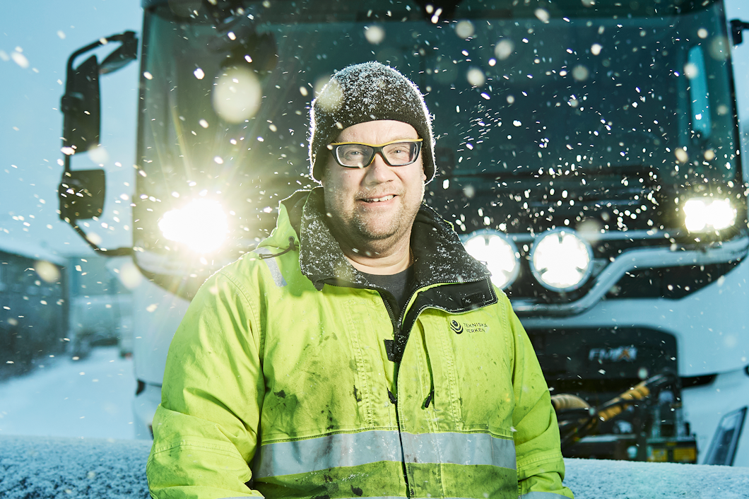 Tony Lövbrand i en högsynlig jacka står framför en snöplog på en dag med snö, med byggnader i bakgrunden.