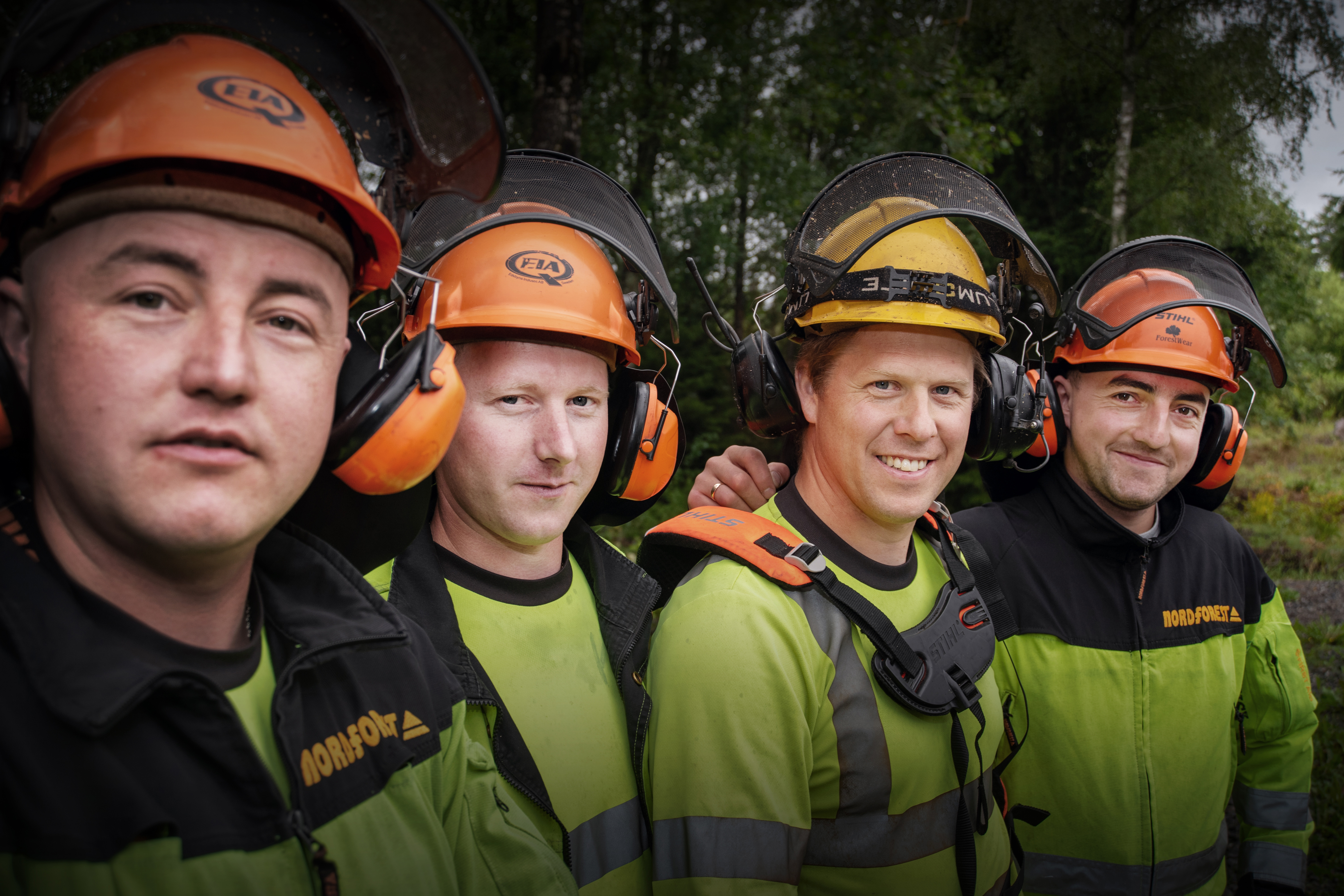 Fyra arbetare i skyddsutrustning, inklusive hjälmar och hörselskydd, står utomhus med träd i bakgrunden.