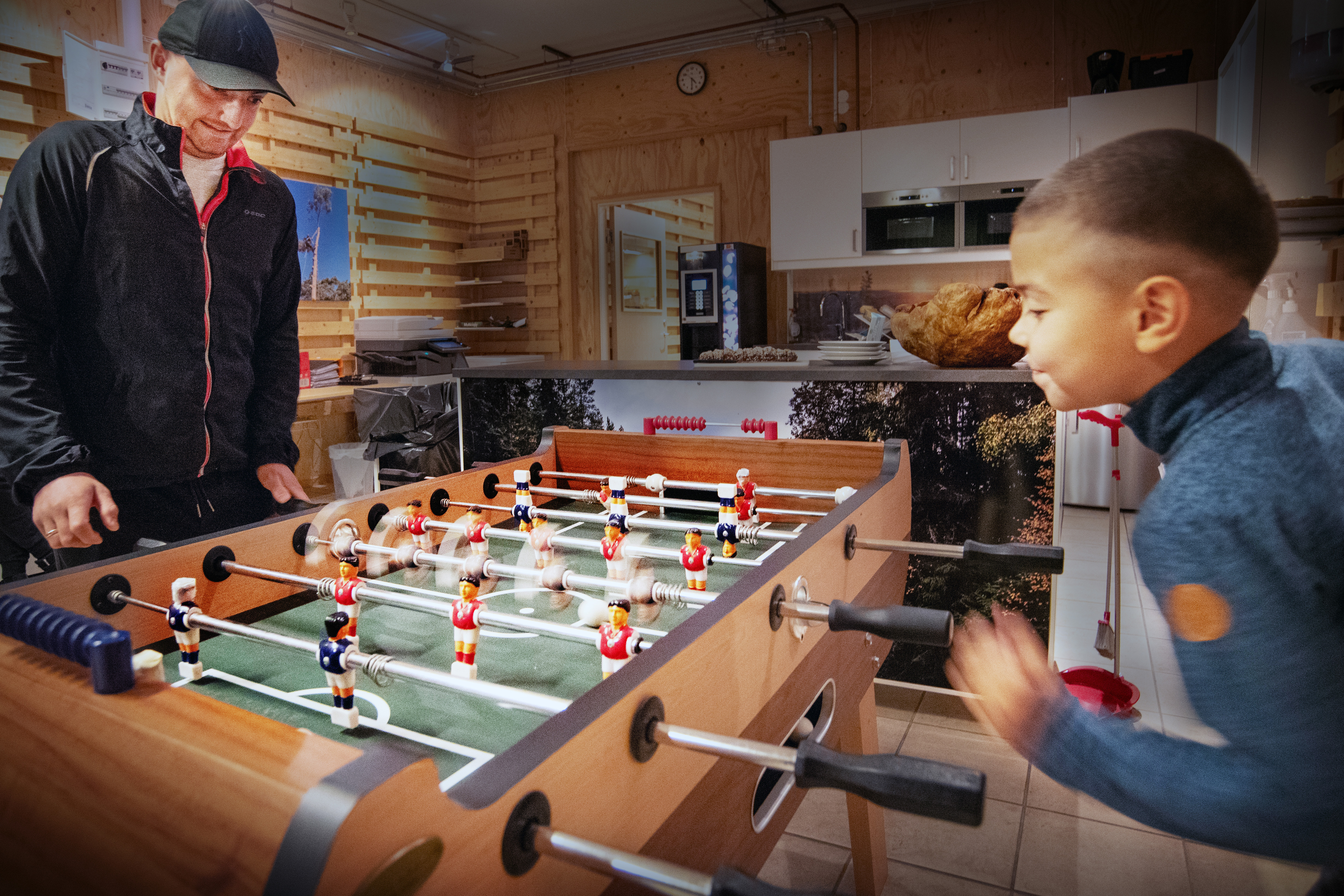 Två personer spelar foosball i ett rum med träväggar och en köksdel i bakgrunden.