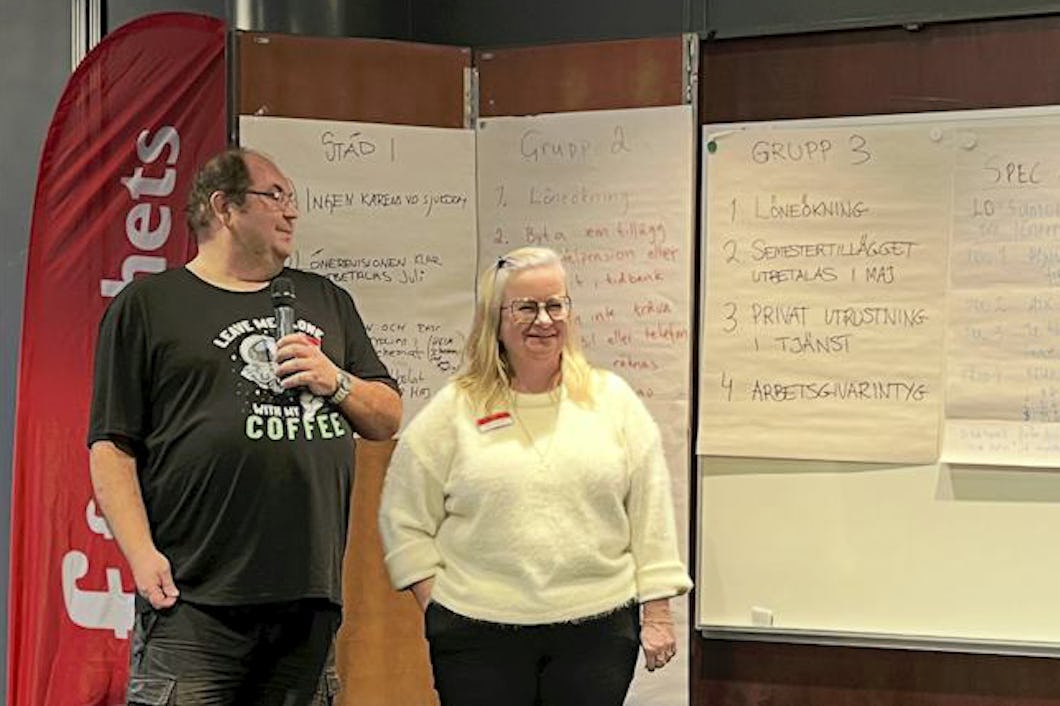En grupp människor står på en liten scen och diskuterar samt skriver på stora pappersark. En person i publiken har färgglatt hår.