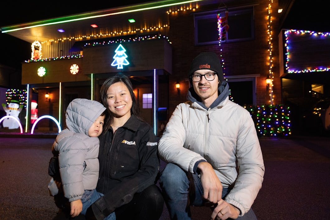 En familj på tre personer står framför ett hus dekorerat med julbelysning och julsaker.