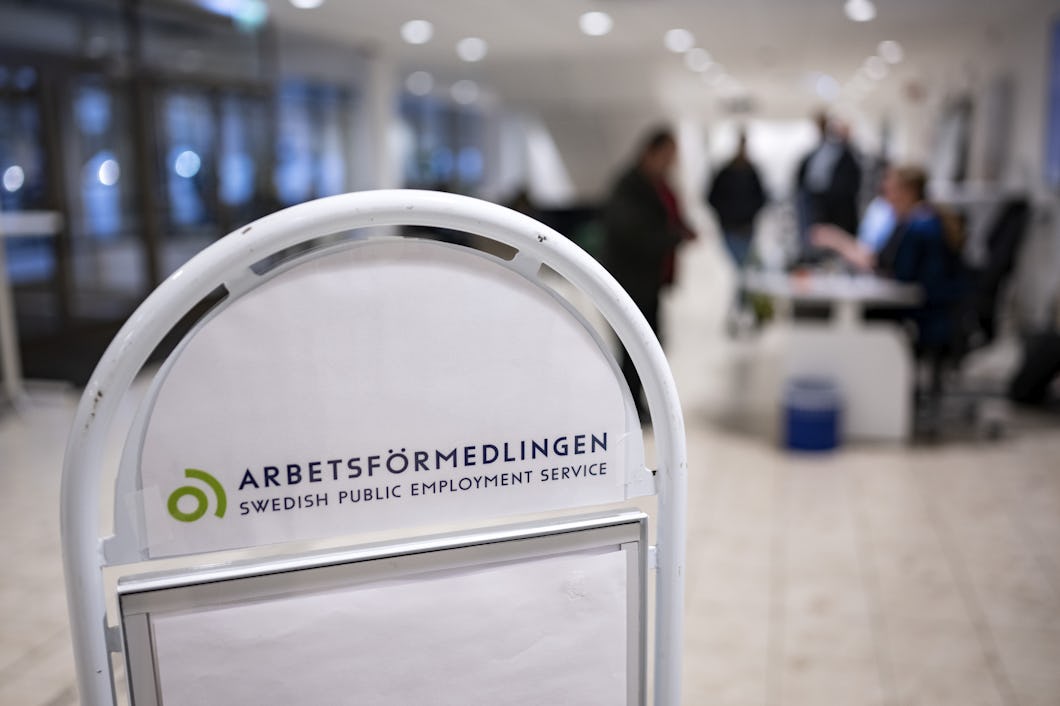 Skylt med texten "Arbetsförmedlingen, Arbetsförmedlingen" i ett modernt kontorsutrymme med människor i bakgrunden.