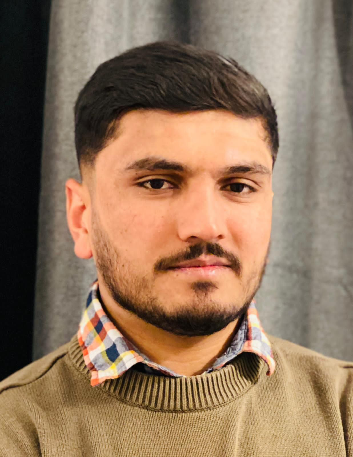 En man med kort svart hår och skägg har på sig en rutig skjorta under en grön tröja. Han står framför ett grått draperi.