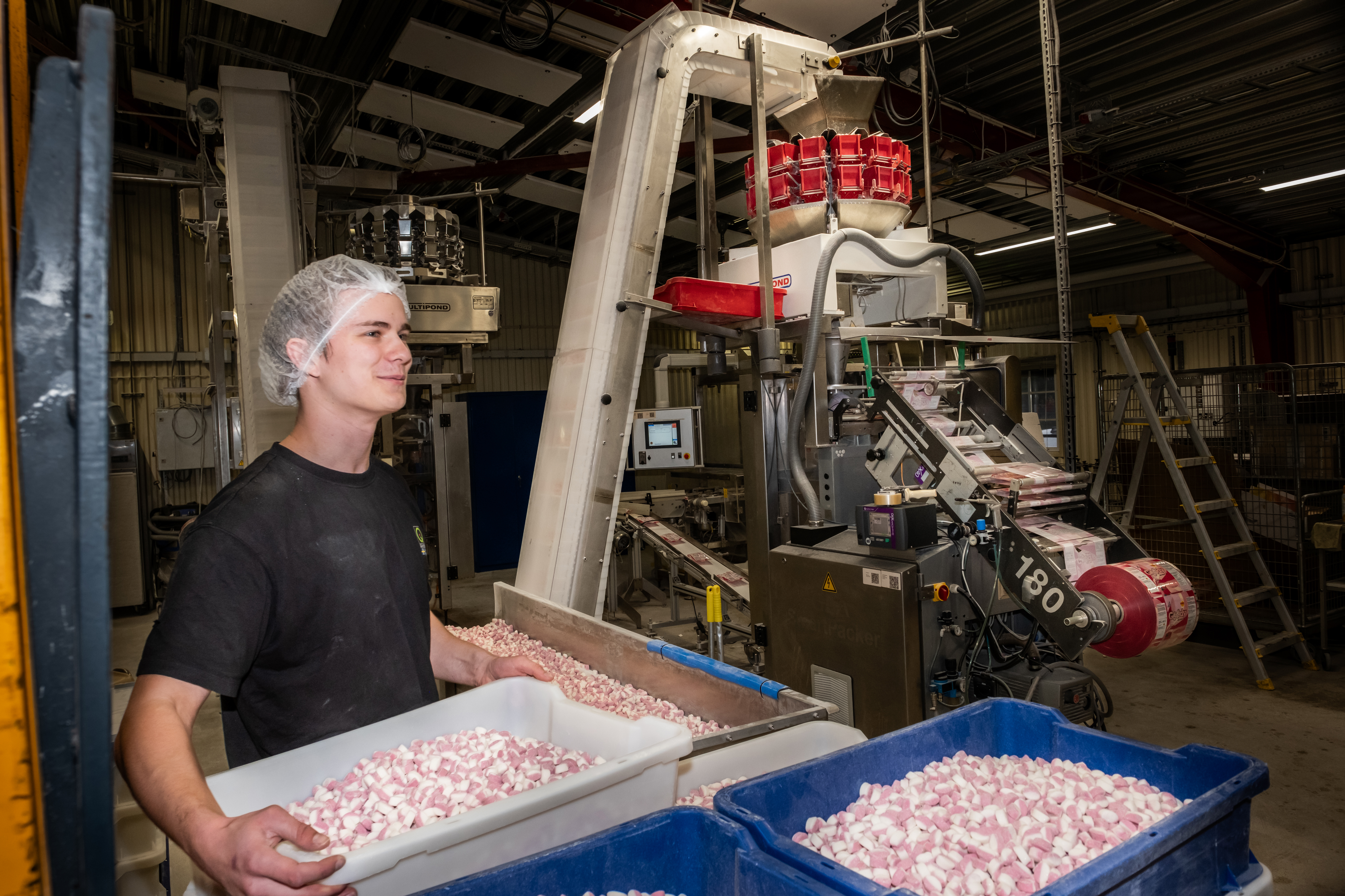 En arbetare med hårnät står i en fabrik och håller i en bricka med randigt godis. I bakgrunden syns maskiner och behållare fyllda med godis.
