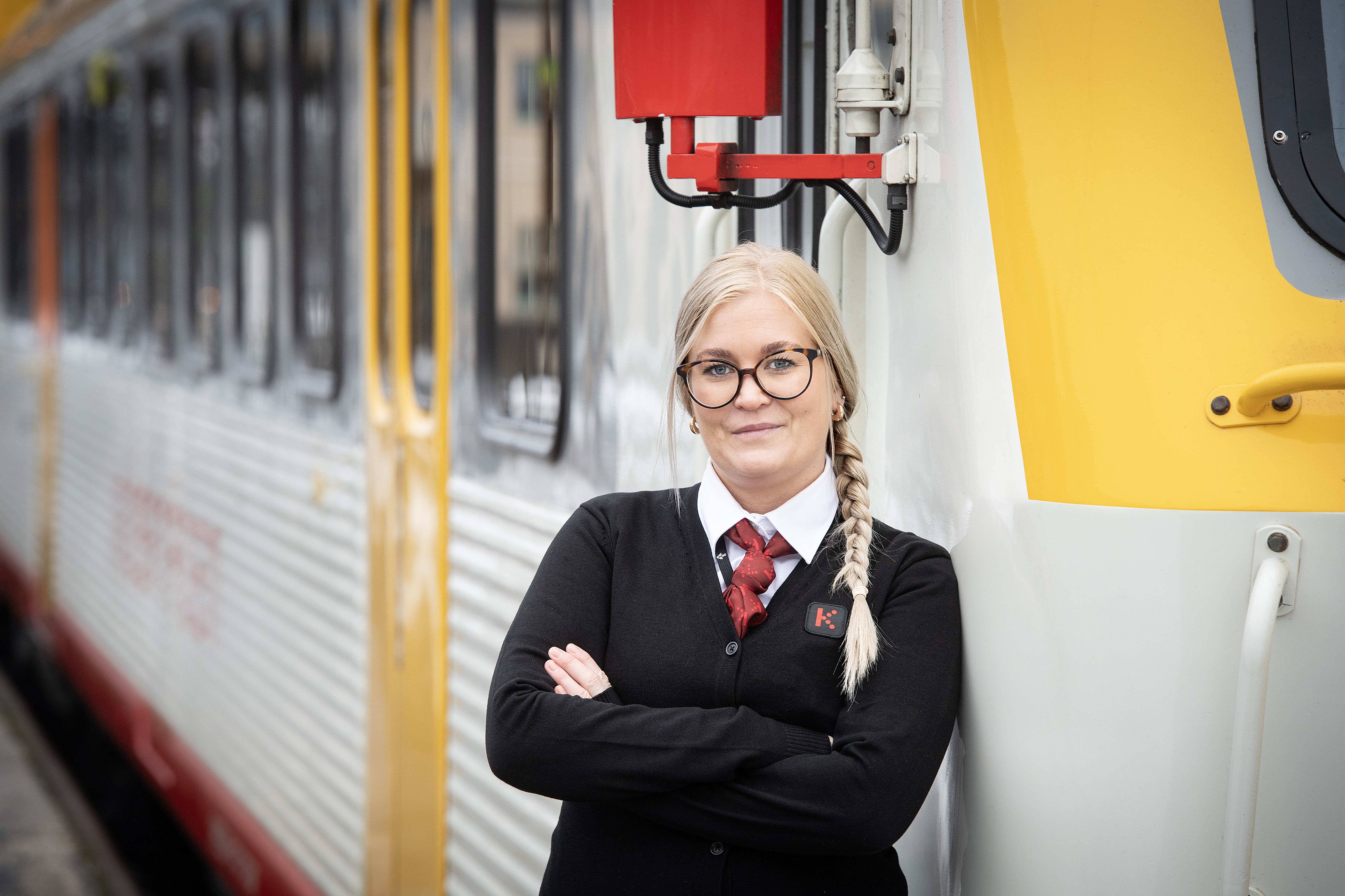 Emelie Lundborg står framför ett Krösatåg, klädd i en svart uniform med röd slips. Armarna är korslagda.