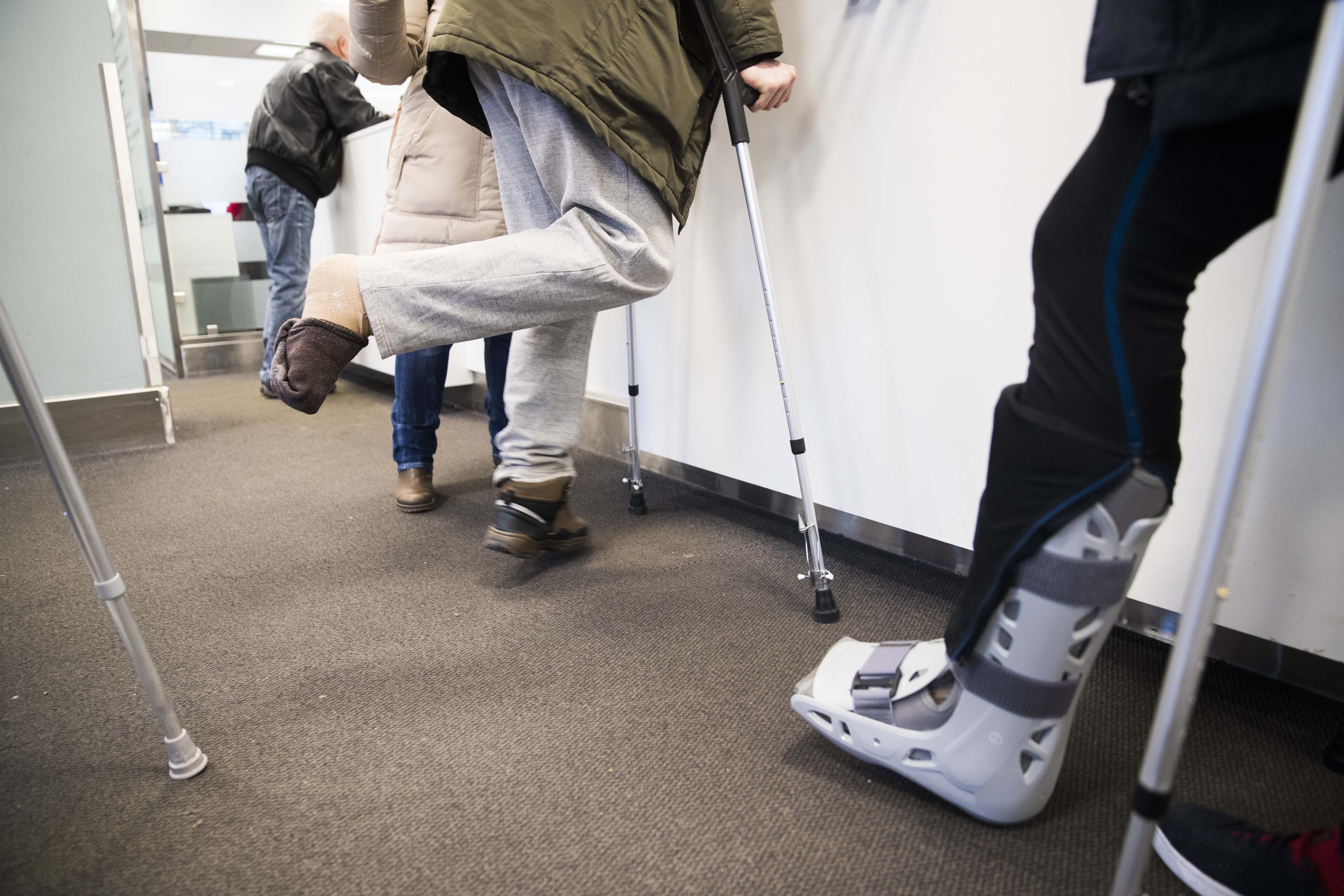 Två personer med benskador går med kryckor. Den ena har en fot inlindad i ett tjockt bandage och den andra bär en ortopedisk stövel. De rör sig framåt i en korridor.