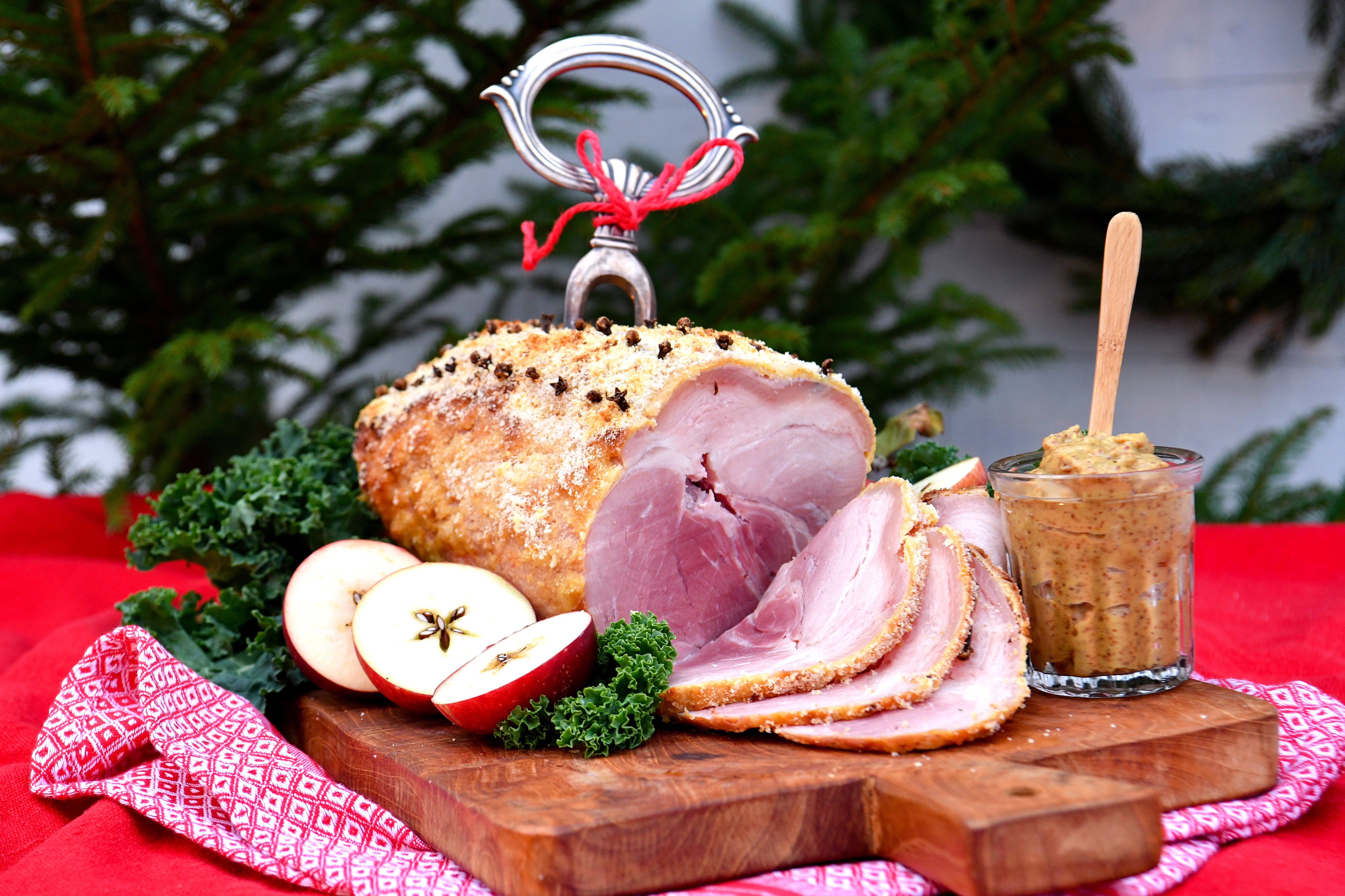 En glaserad skinka garnerad med örter är skivad på en träbräda, med äppelskivor och en burk senap bredvid. En röd duk ligger i bakgrunden.