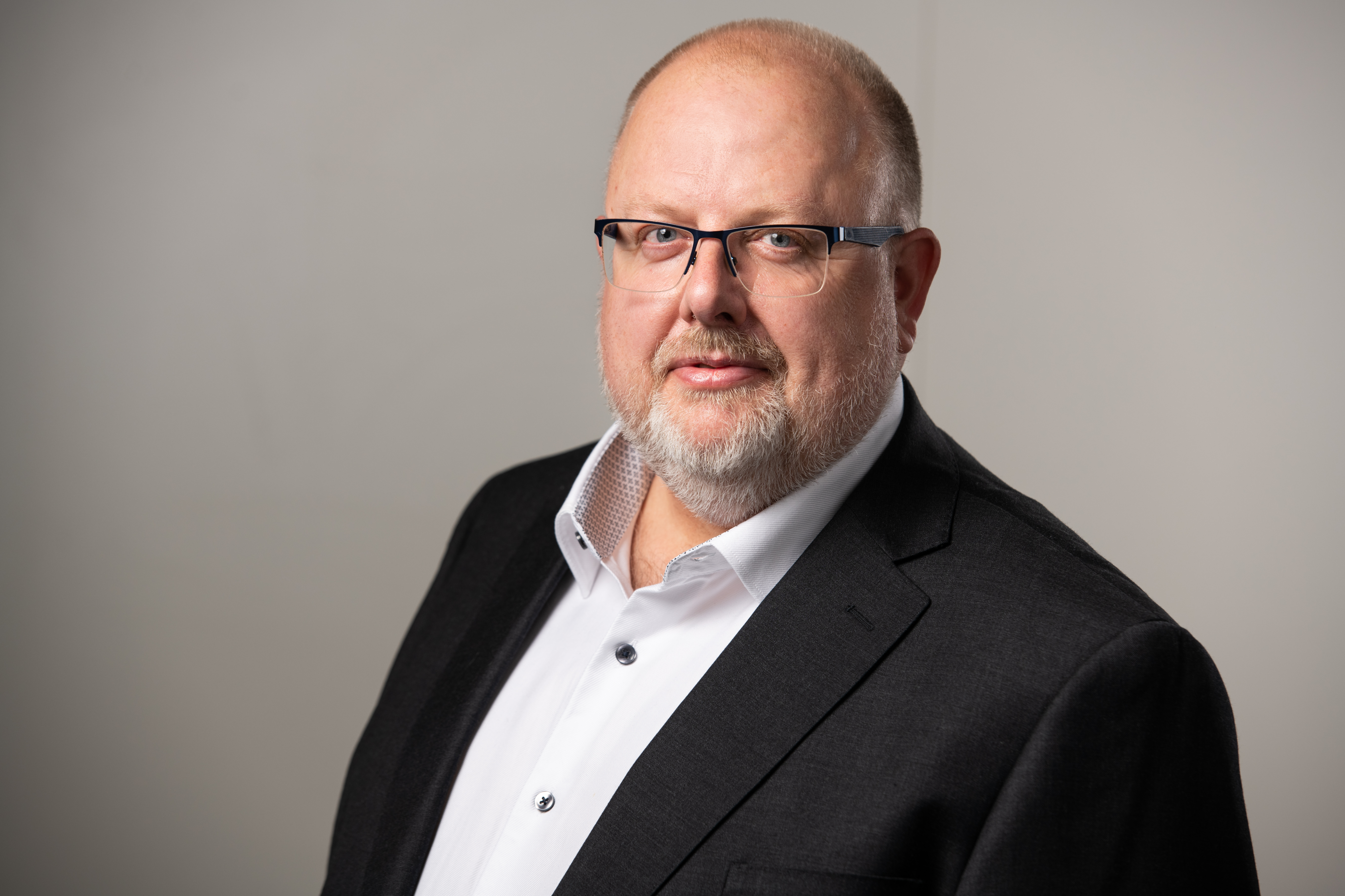 Man med glasögon och skägg bär en svart kostym och vit skjorta, står mot en enkel grå bakgrund.