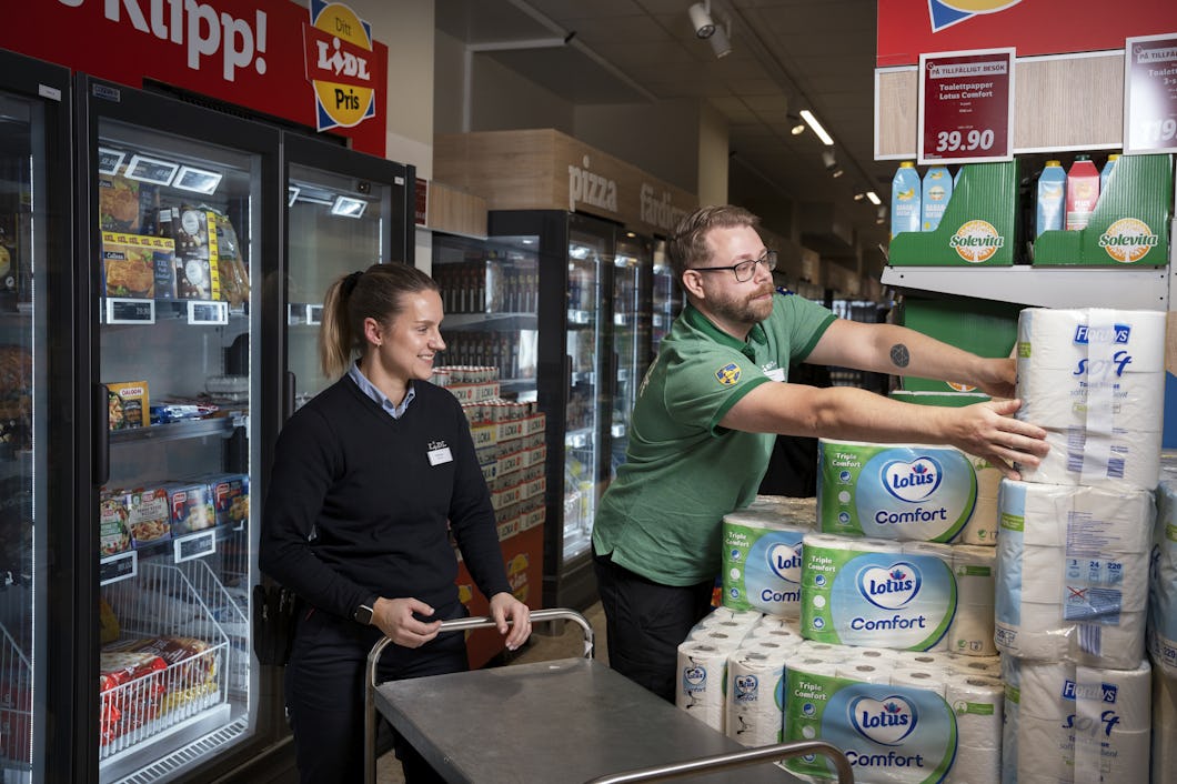Två butiksanställda som organiserar förpackningar med toalettpapper nära kylda varor i en gång i mataffären.