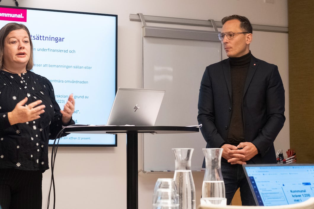 Två personer befinner sig i en kontorsmiljö och håller en presentation. En person står nära en skärm som visar text, medan den andra personen lyssnar. På bordet finns flera glas och en bärbar dator.