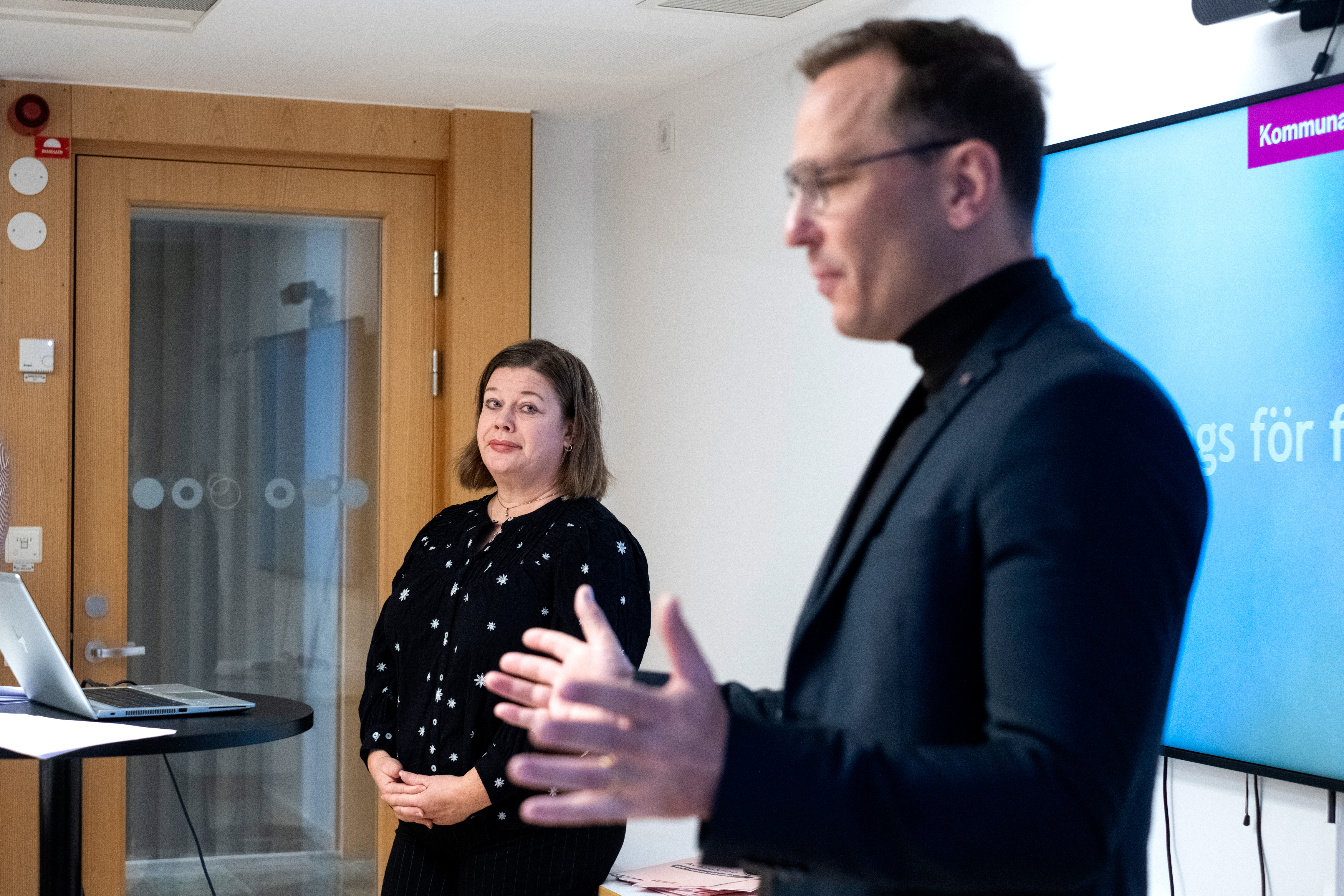 En man håller en presentation framför en stor skärm medan en kvinna står vid en bärbar dator och lyssnar uppmärksamt.