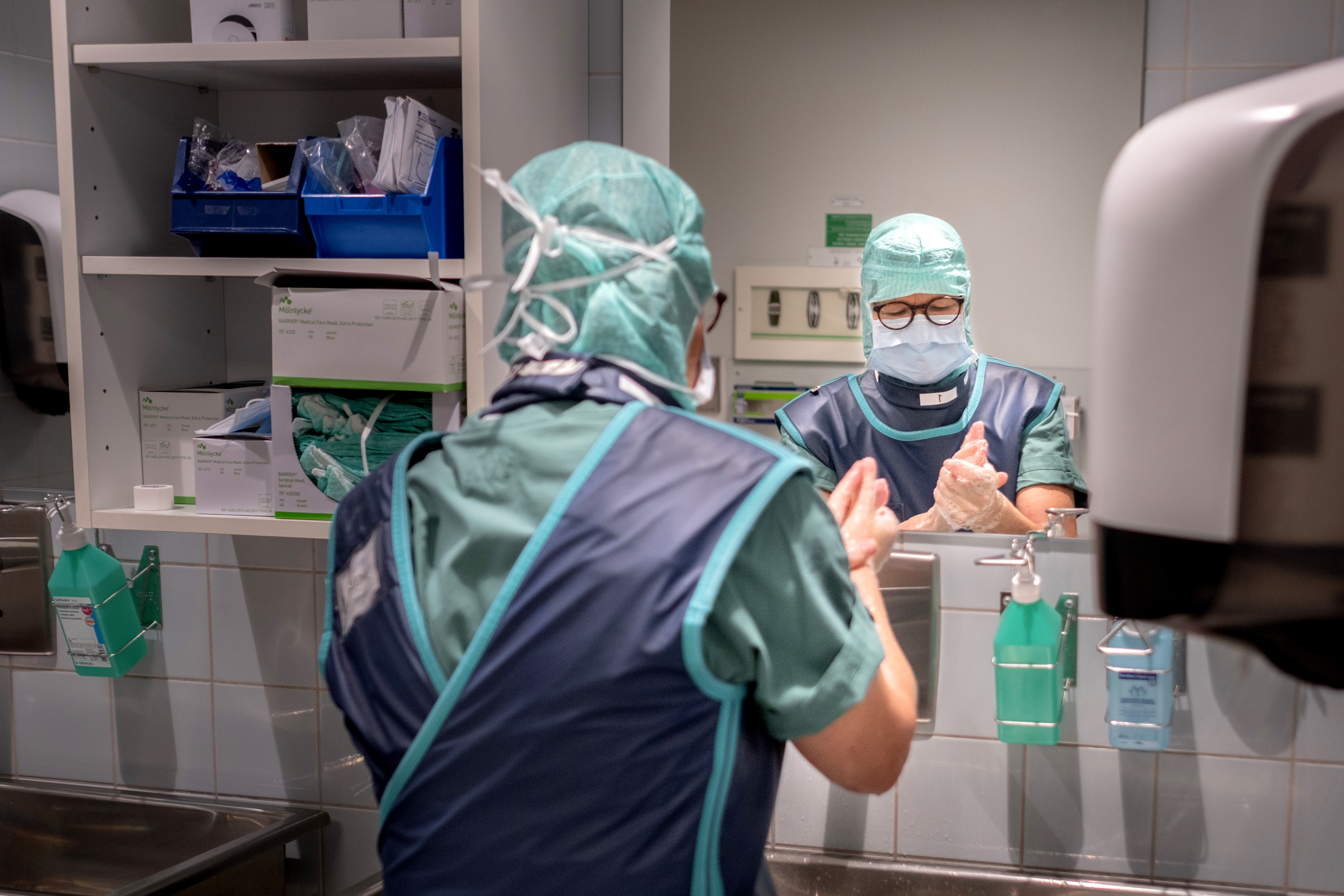 En vårdpersonal iklädd sjukhuskläder och skyddsutrustning tvättar händer i en sjukhusmiljö.