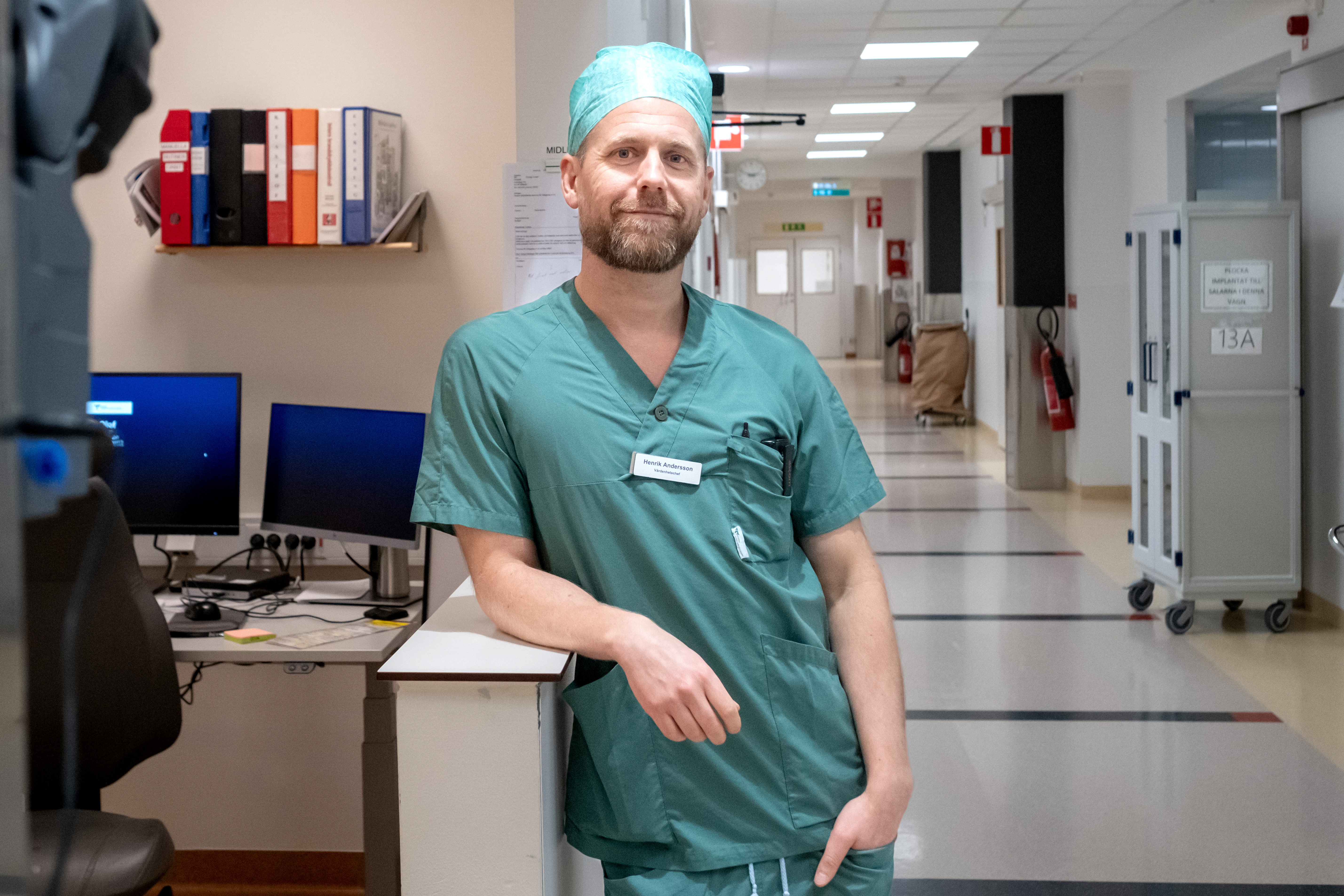 En person i gröna sjukhuskläder och mössa lutar sig mot ett skrivbord i en sjukhuskorridor. Två datorskärmar och kontorsmaterial syns i bakgrunden.