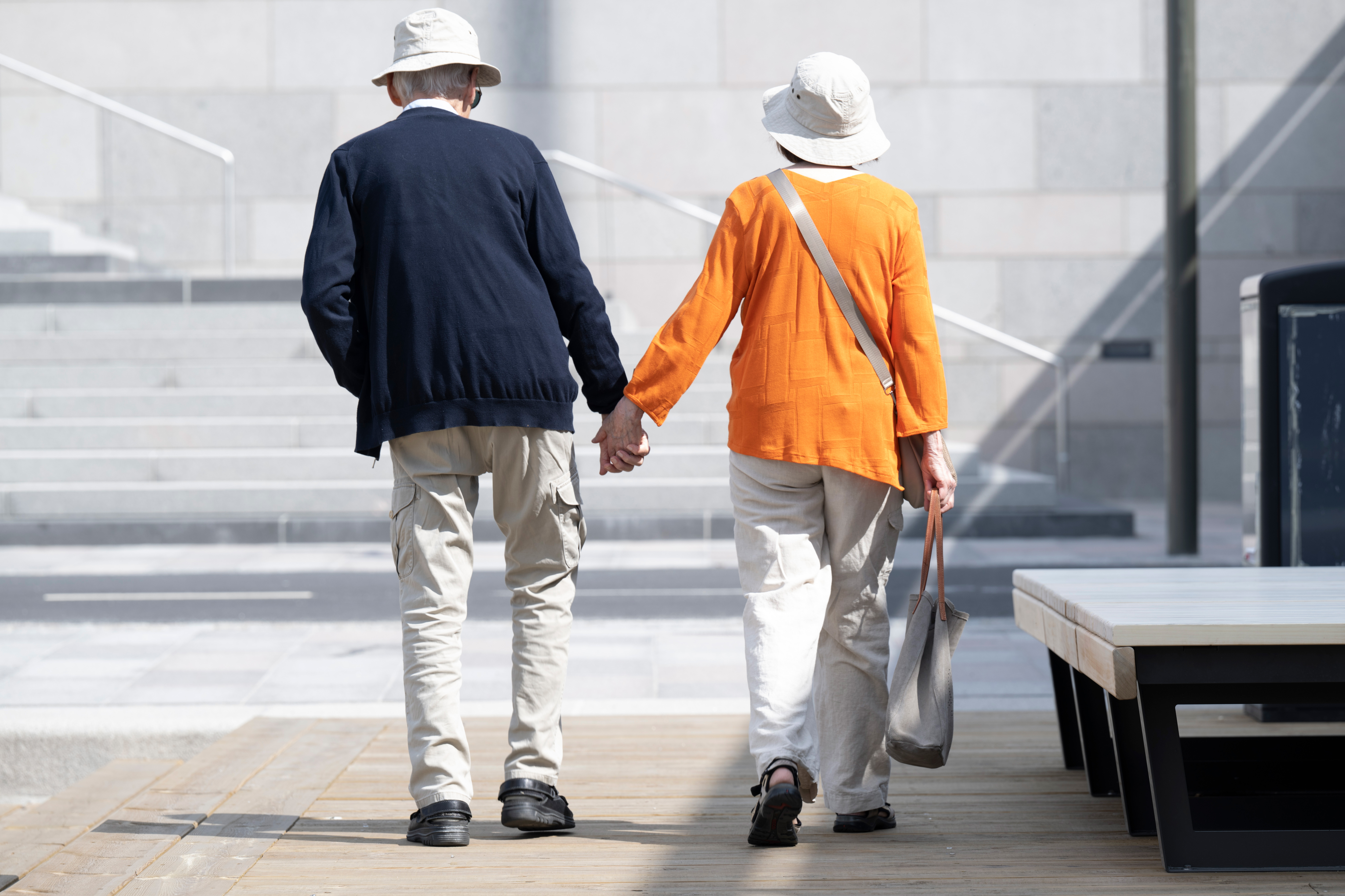 Två äldre personer går hand i hand på en trästig. Den ena bär en hatt och en orange skjorta, den andra har på sig en hatt och en svart jacka.