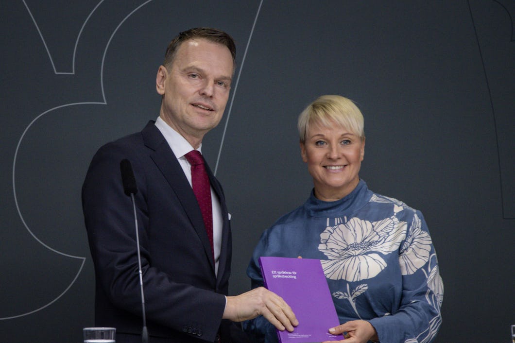 Peter Danielsson och Anna Tenje i formella kläder ler och håller tillsammans i ett lila häfte framför en mörk bakgrund.