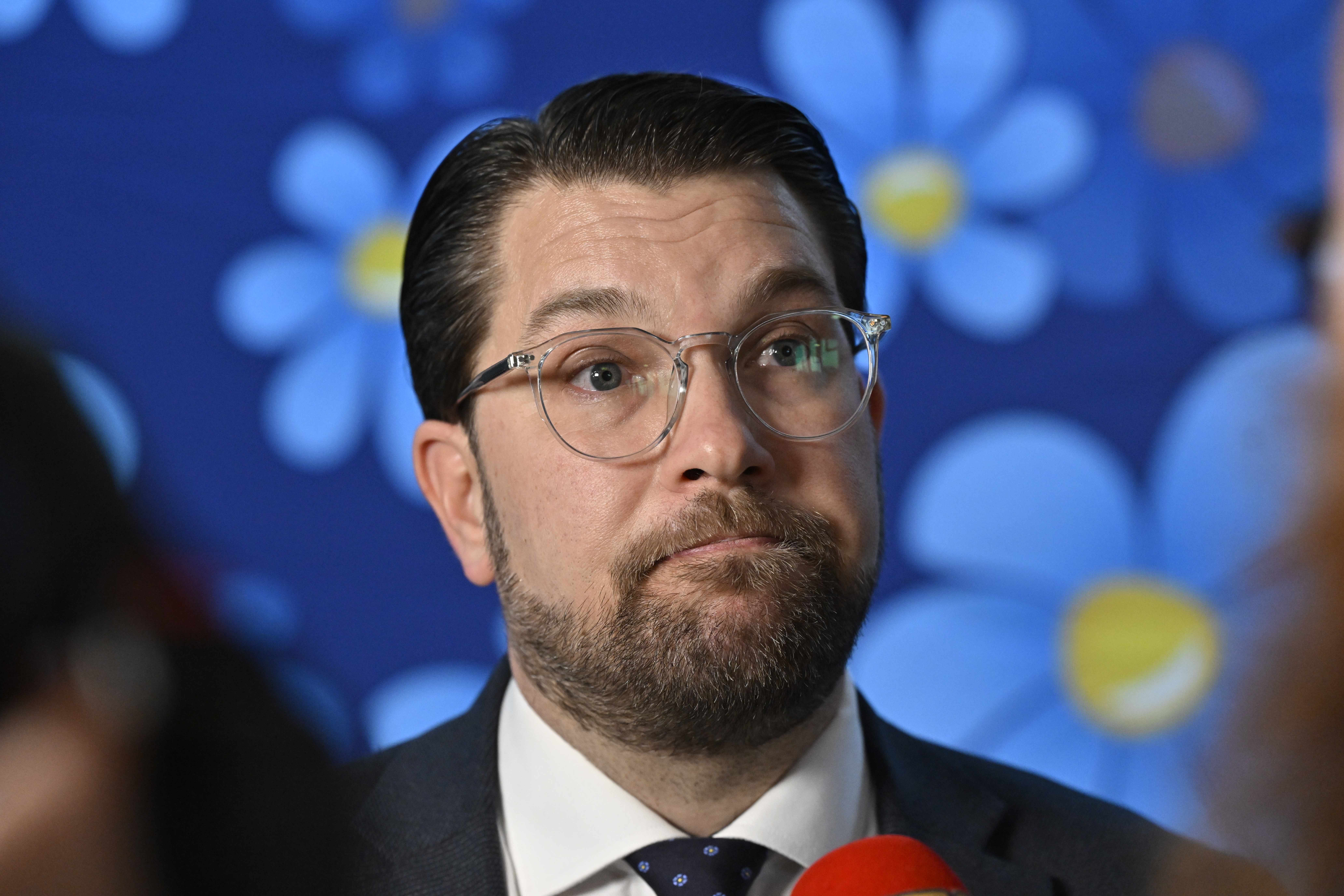Jimmie Åkesson med glasögon och en kostym står framför en blå blomsterbakgrund och håller i en röd mikrofon.
