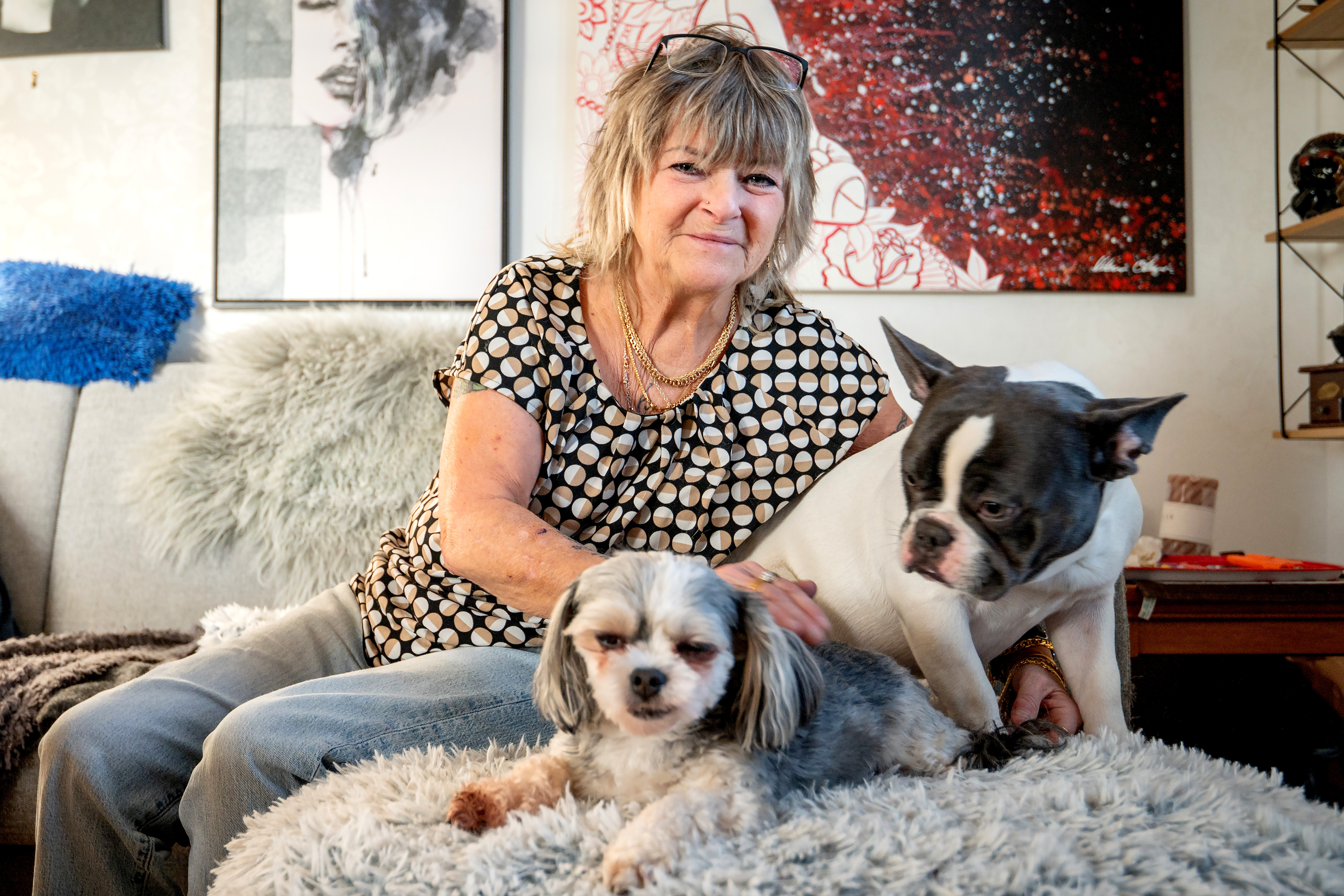 En kvinna sitter på en soffa med en liten hund i knäet och en större hund bredvid sig. Bakom henne syns inramad konst på väggen.
