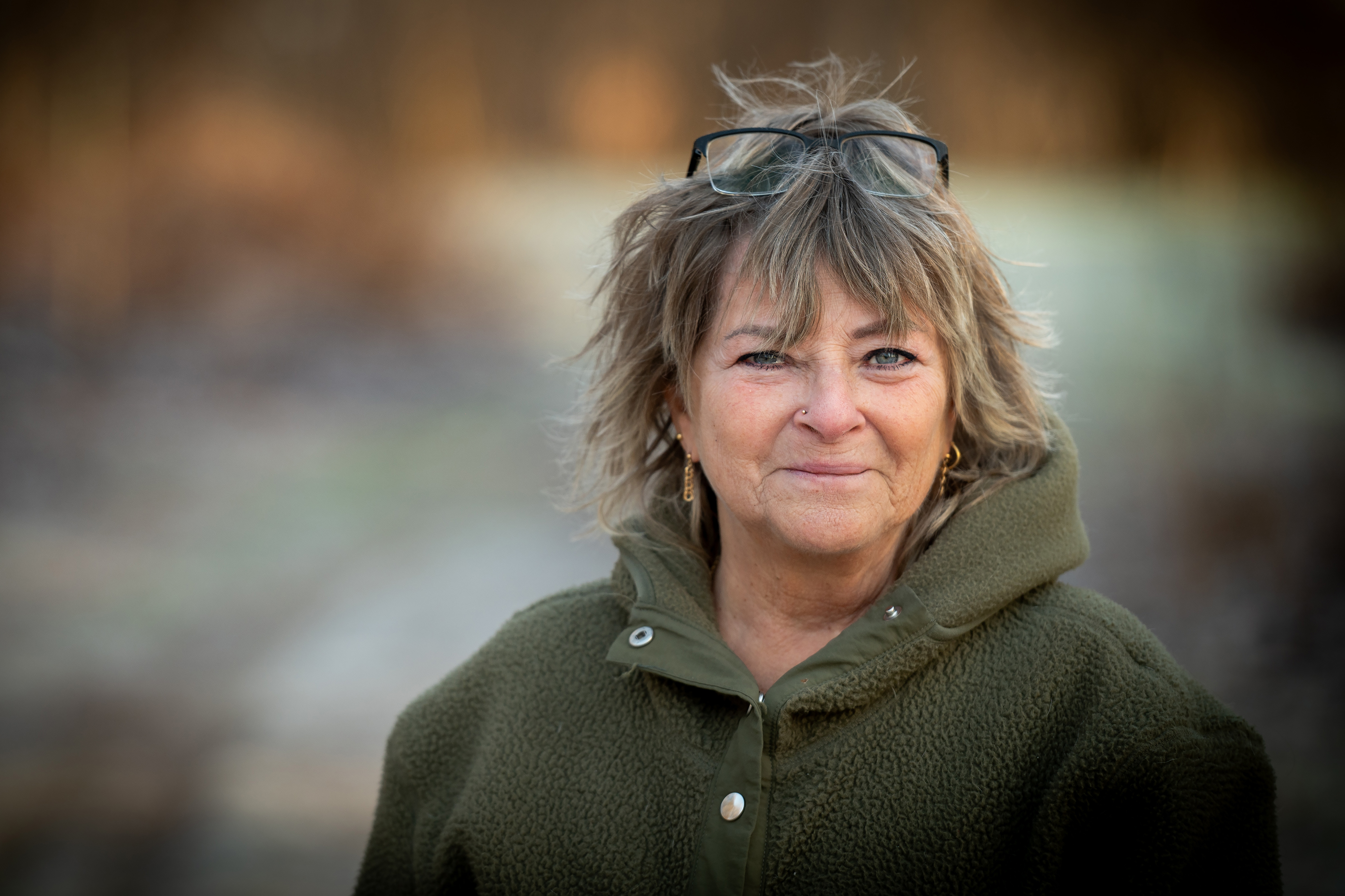 En person med kort hår, iklädd en grön jacka och har glasögon på huvudet, står utomhus och tittar in i kameran. Bakgrunden är något suddig.