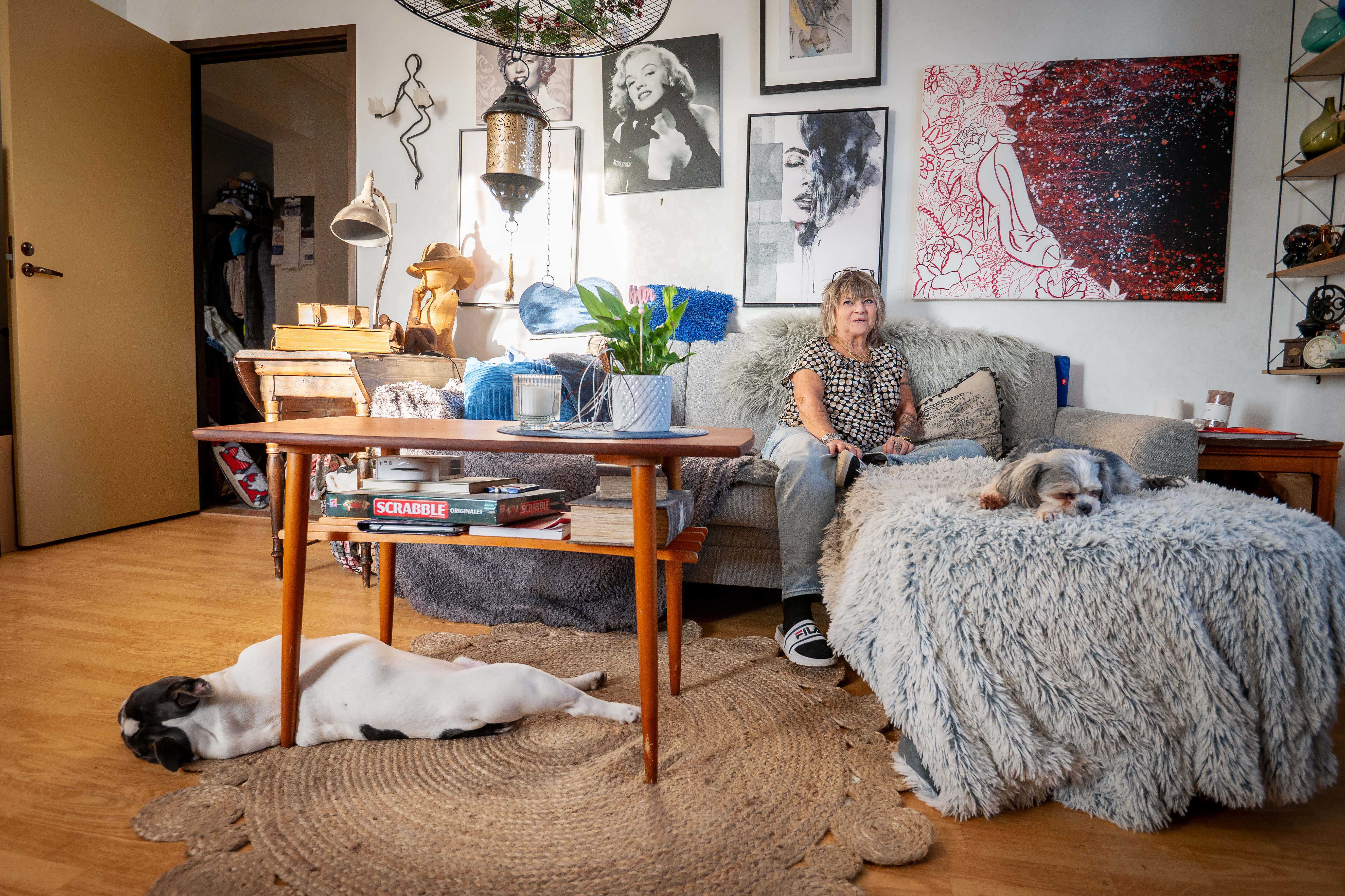 En kvinna sitter på en soffa i ett vardagsrum med två sovande hundar. Runt om henne finns konstverk och dekorationer.