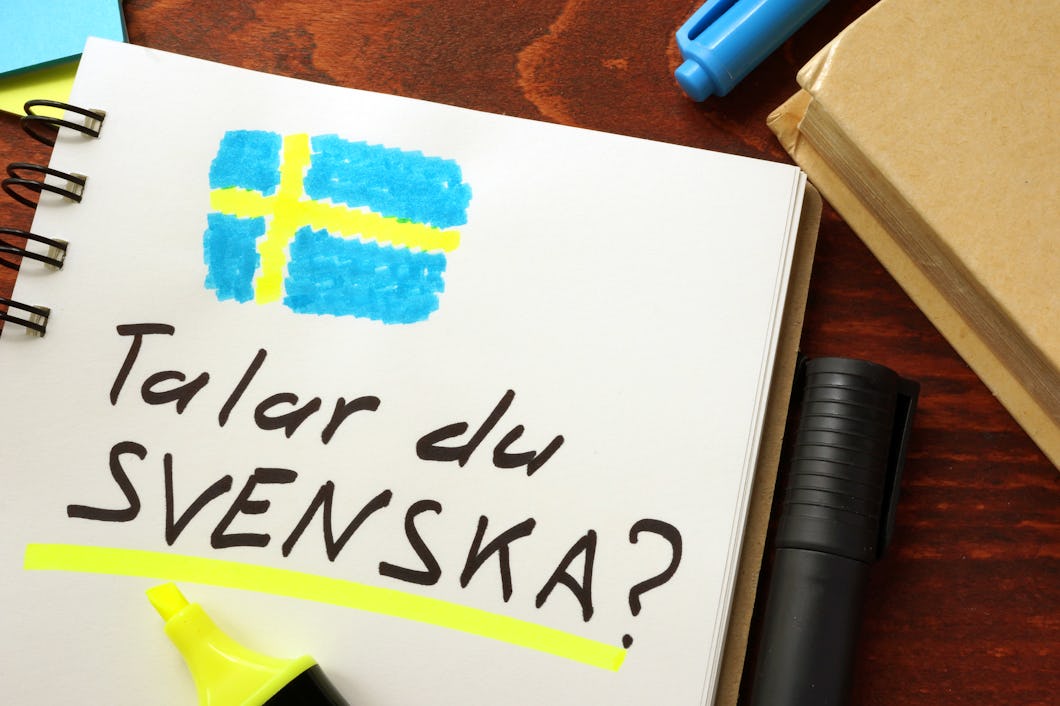 En anteckningsbok med texten "Talar du svenska?". En handritad svensk flagga och olika skrivbordstillbehör syns.