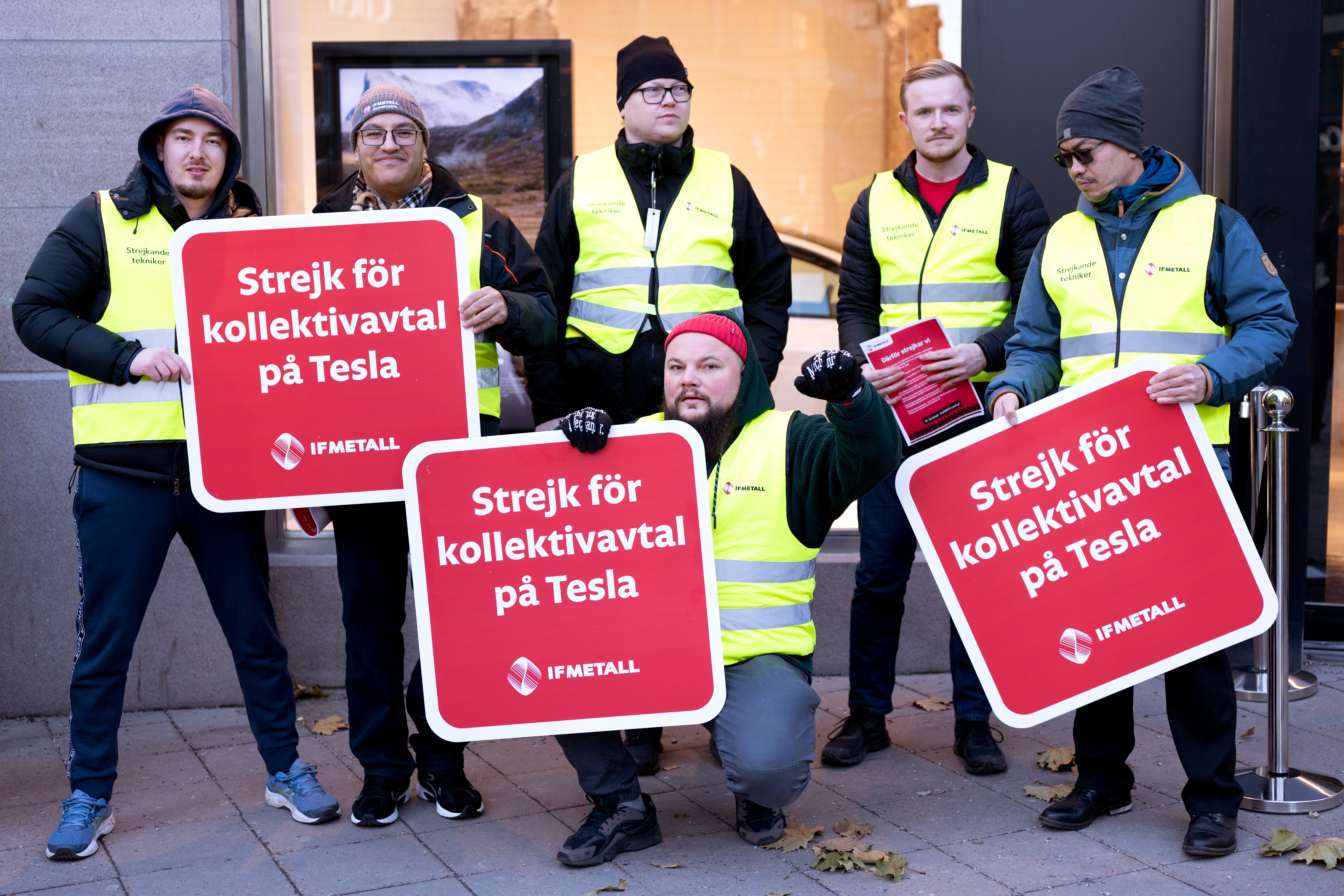 Sju personer i gula västar står på en trottoar och håller i skyltar med texten "Strejk för kollektivavtal på Tesla" på svenska.