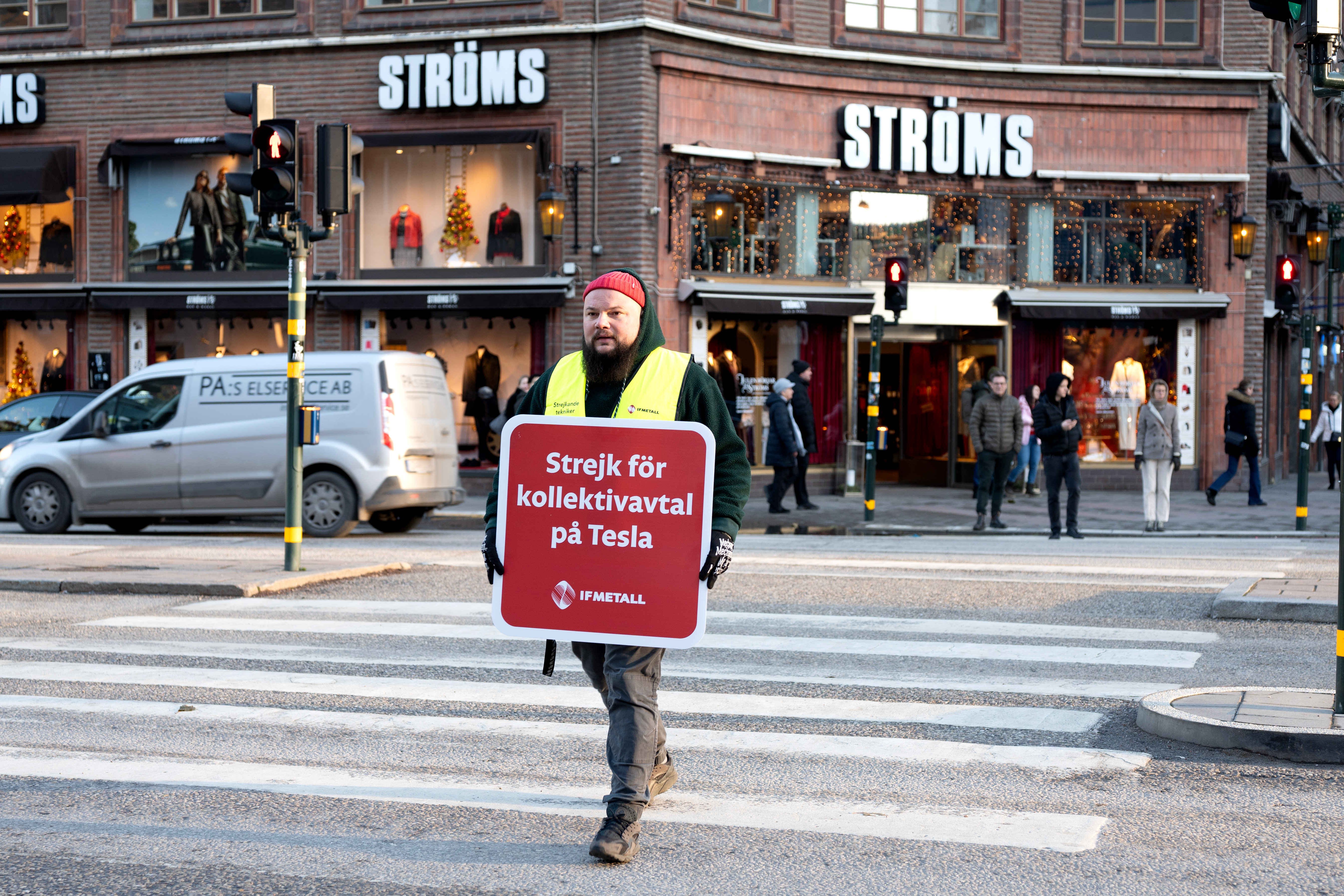 En person håller en skylt med texten "Strejk för kollektivavtal på Tesla" och går över en gata. Människor syns i bakgrunden tillsammans med en butik som heter Ströms.
