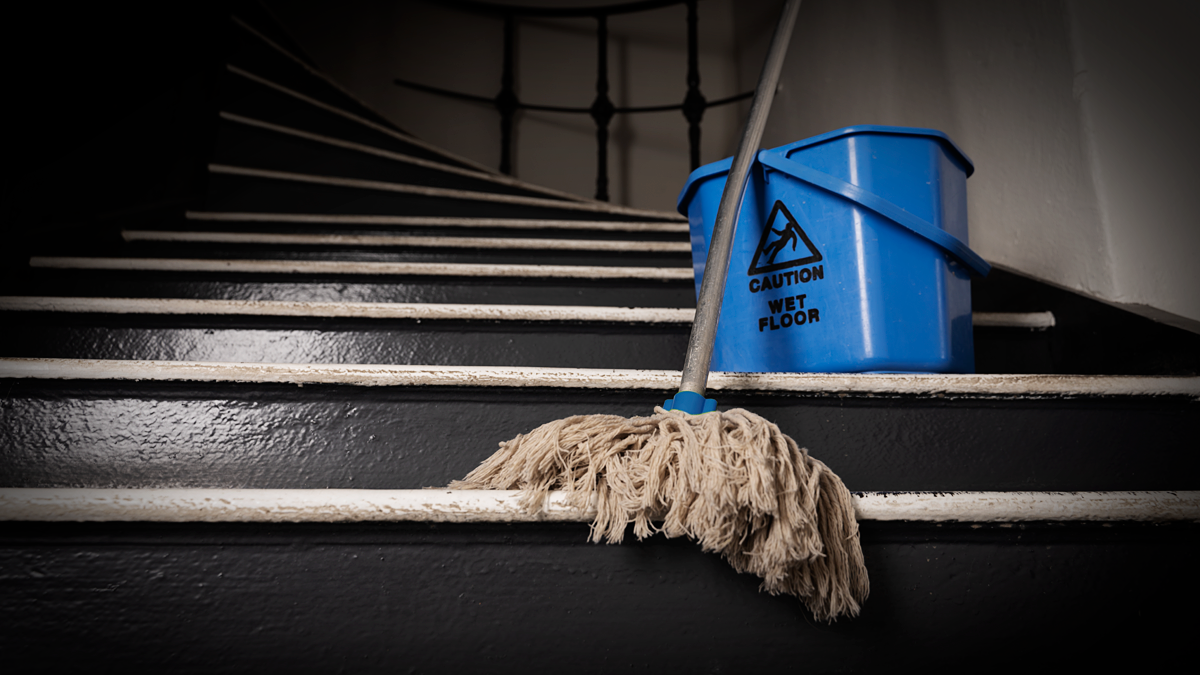 En mopp lutar mot en gammal trappa, med en blå hink märkt "Varning Vått Golv" placerad på ett av trappstegen.