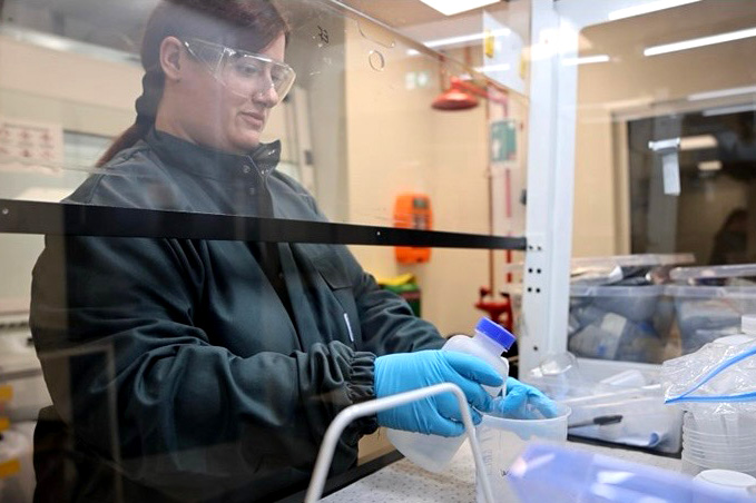 En kvinna med skyddsglasögon och handskar häller vätska i en behållare i en laboratoriemiljö.