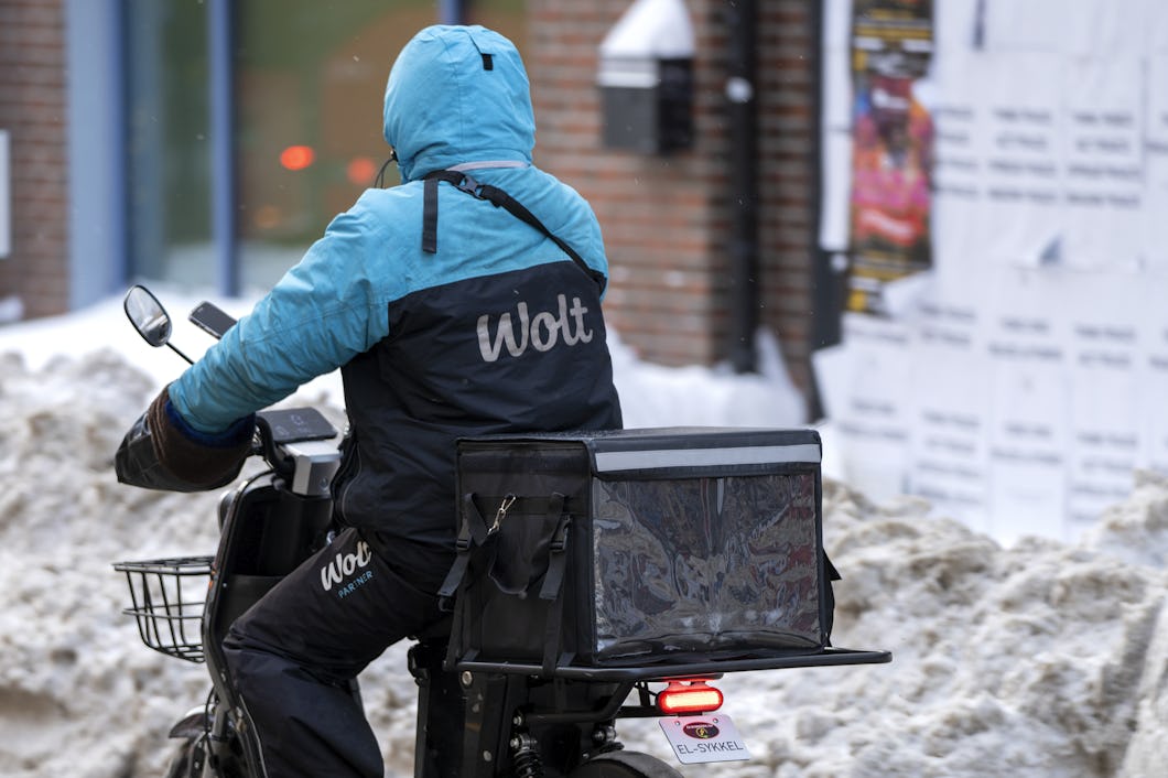 En budbärare med en blå Wolt-jacka cyklar på en elcykel genom en snötäckt gata.