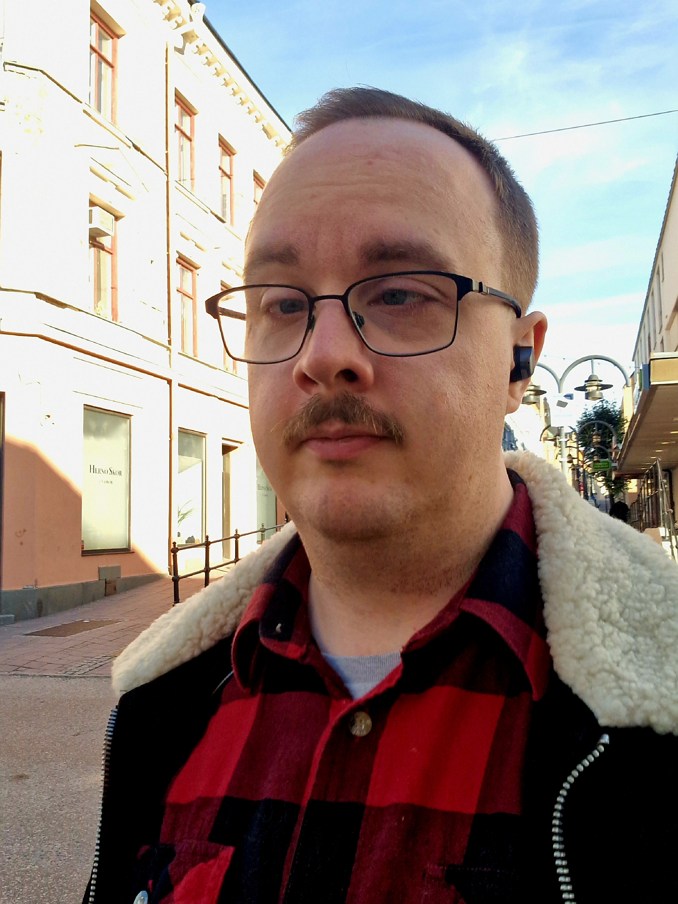 undersköterskan Emil Olsson med glasögon och mustasch bär en röd rutig skjorta och jacka. Personen står på en upplyst gata med byggnader i bakgrunden.