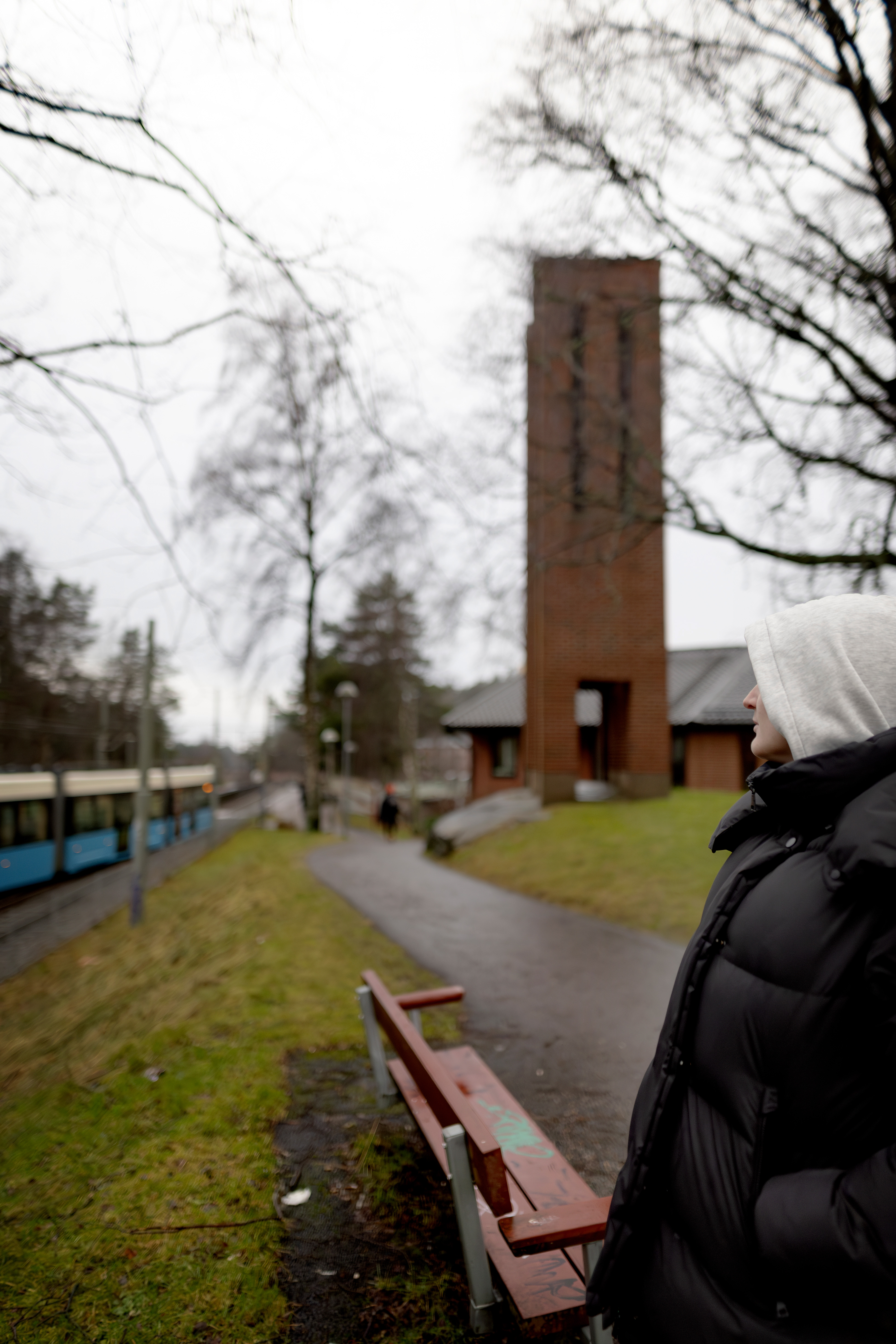 En person iklädd en jacka med huva står nära en stig med en röd bänk. I bakgrunden syns en byggnad med ett högt tegeltorn. En spårvagn kan ses till vänster under en mulen dag.