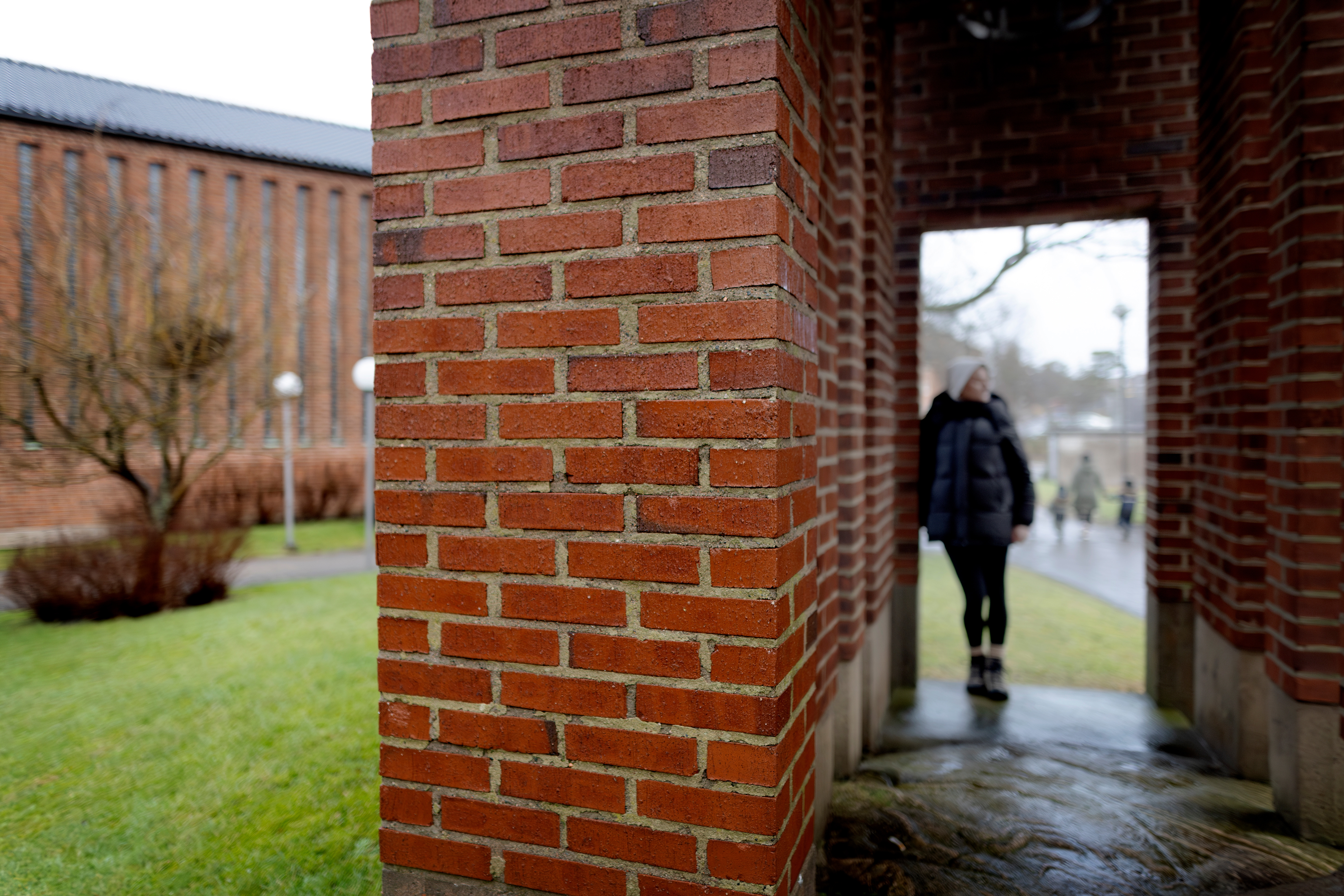 En person i svart rock går genom en tegelvalv en fuktig dag, med en byggnad och ett kalt träd i bakgrunden.