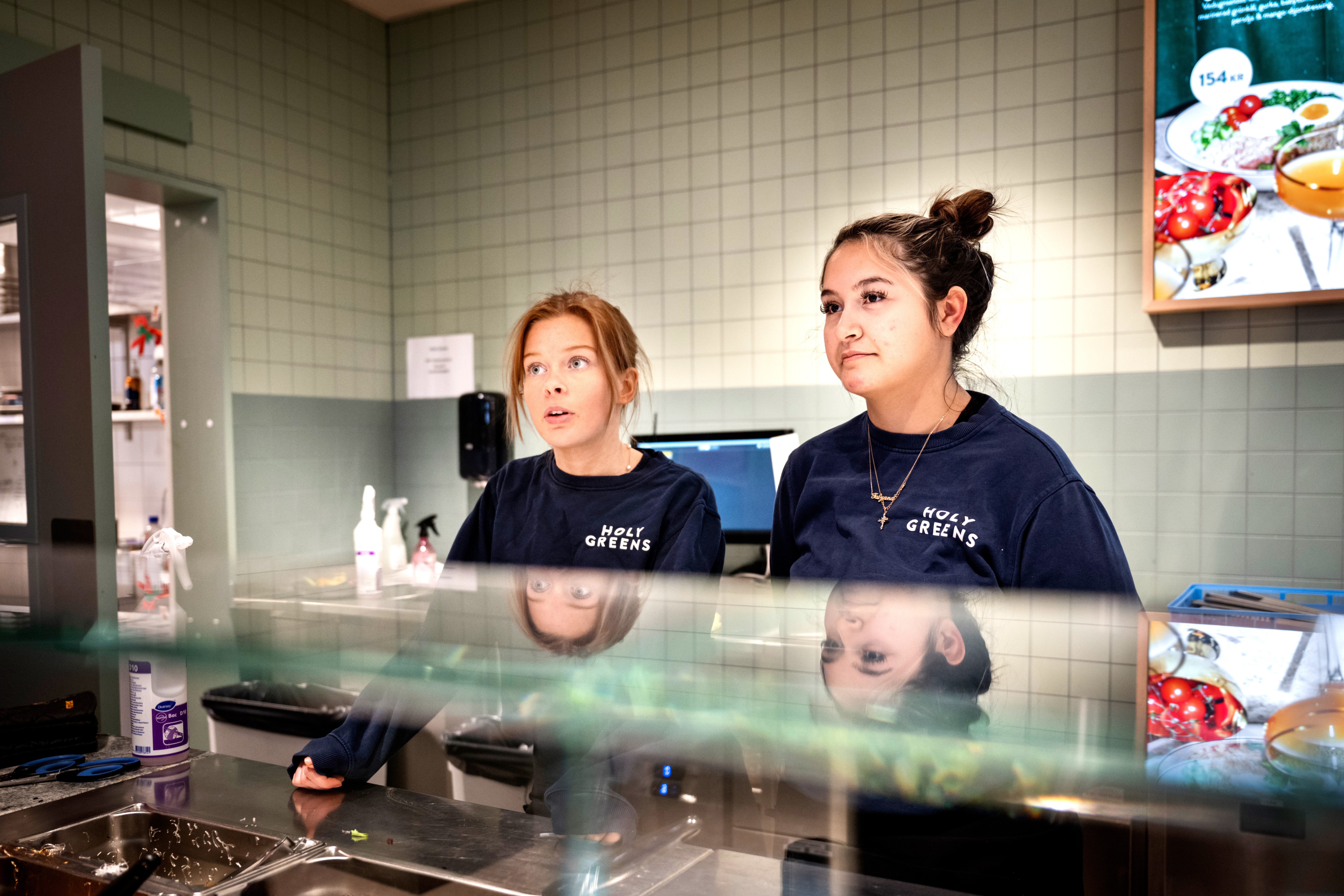 Två anställda står bakom en disk i en matbutik, klädda i mörkblå skjortor med texten "HOLY GREENS".
