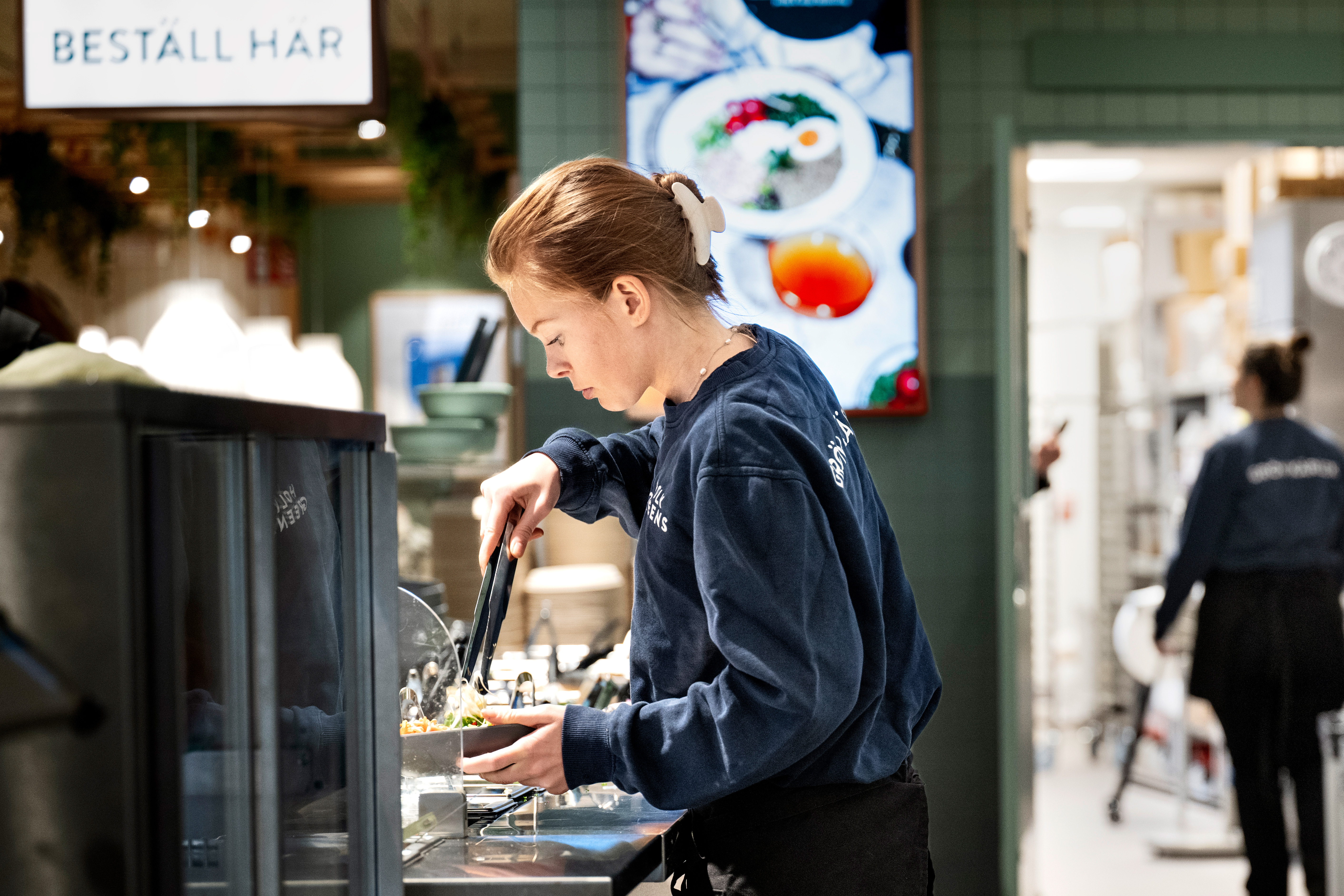 Person som förbereder mat bakom en disk i en restaurang, iklädd blå skjorta. En meny är synlig i bakgrunden.