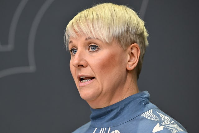 En person med kort, ljust blont hår pratar. Personen har på sig en blåvit mönstrad topp. Bakgrunden är grå.