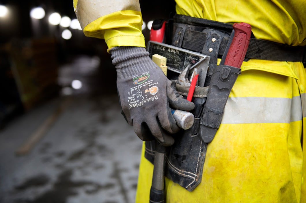 Person i gul uniform och handskar som håller en hammare i ett verktygsbälte.