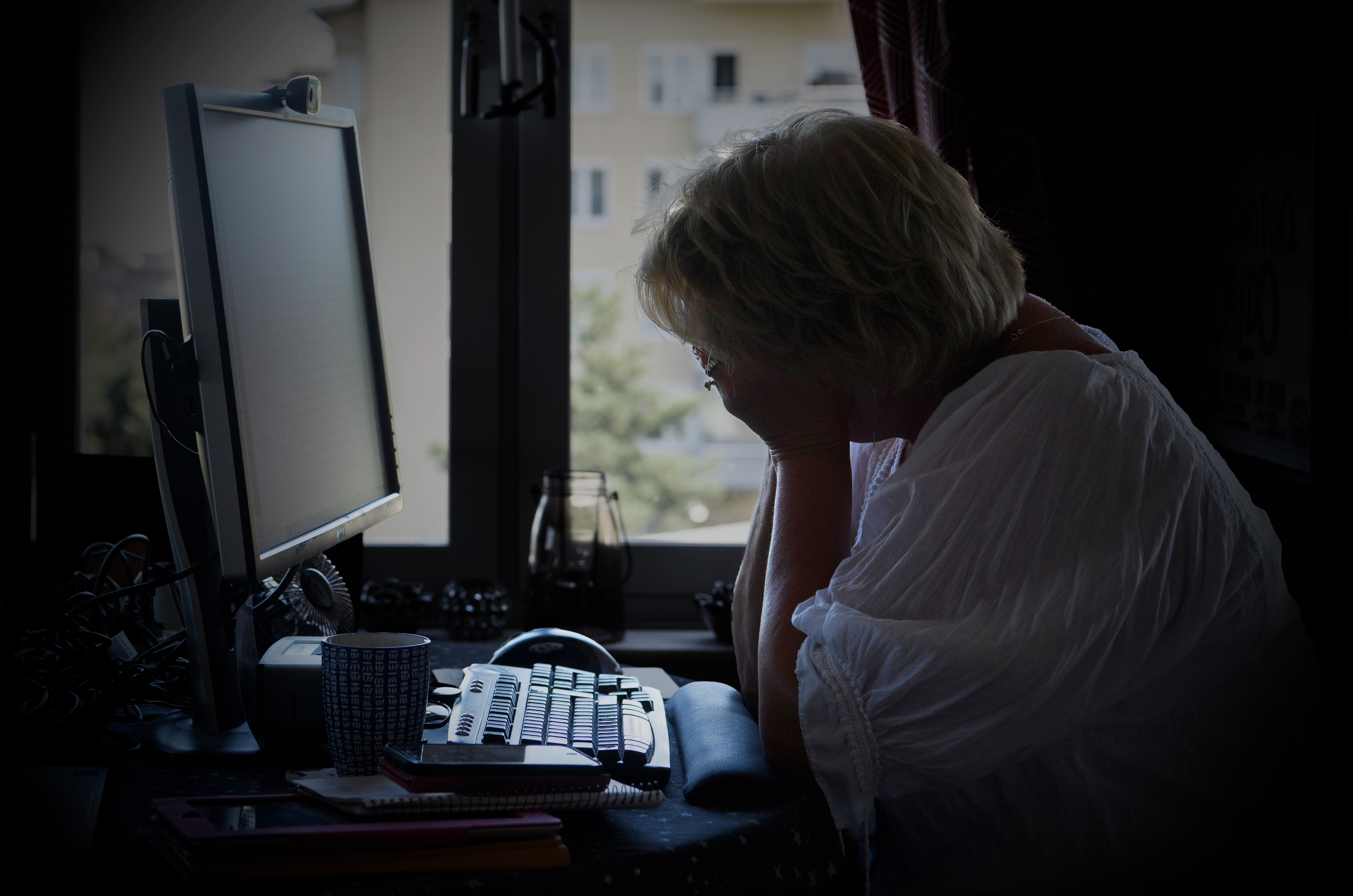 En person sitter vid ett skrivbord med en dator, vill huvudet i händerna, nära ett fönster med byggnader synliga utanför.